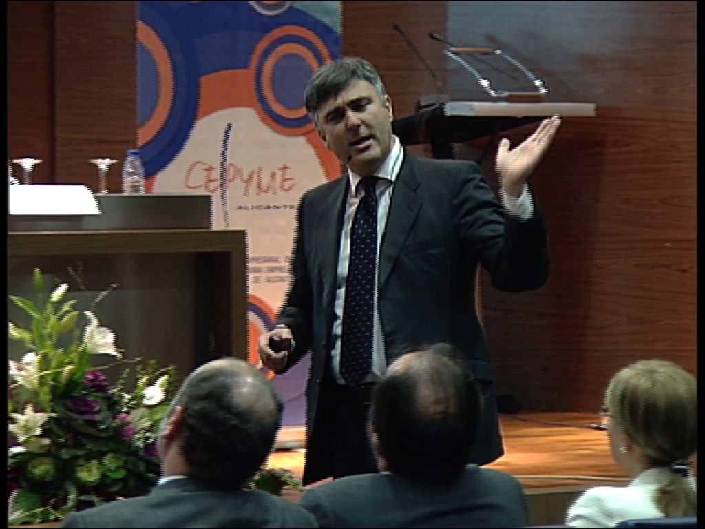 At the bottom of the picture, we see two men and a woman are sitting on the chairs. In front of...