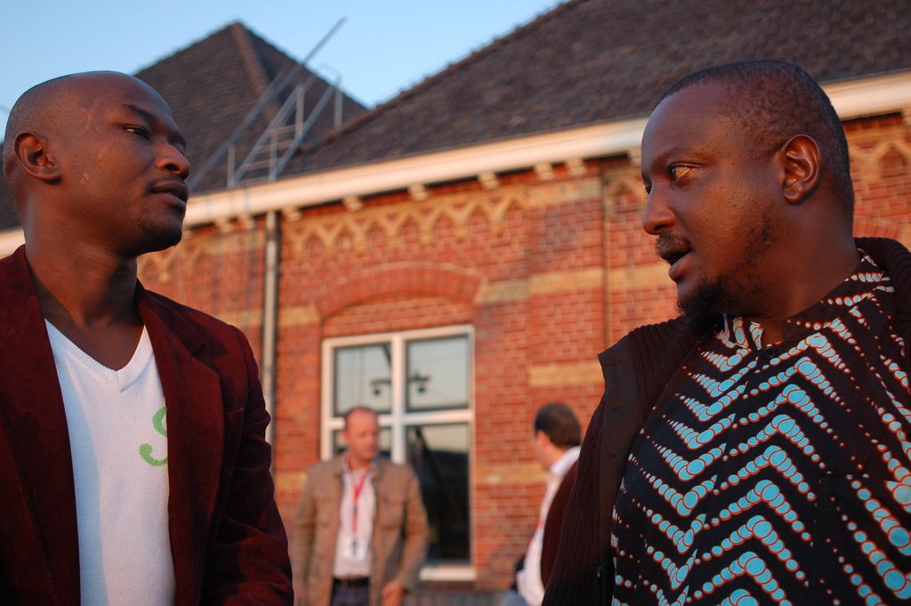 In this picture there are two African boys standing and discussing something. Behind there is a red...