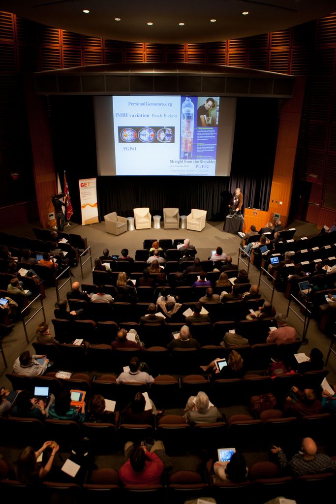 In this image we can see an auditorium. There are many people sitting on the chairs and holding...