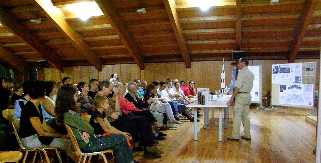 In this image there are group of people sitting in the hall and listening to the person who is...