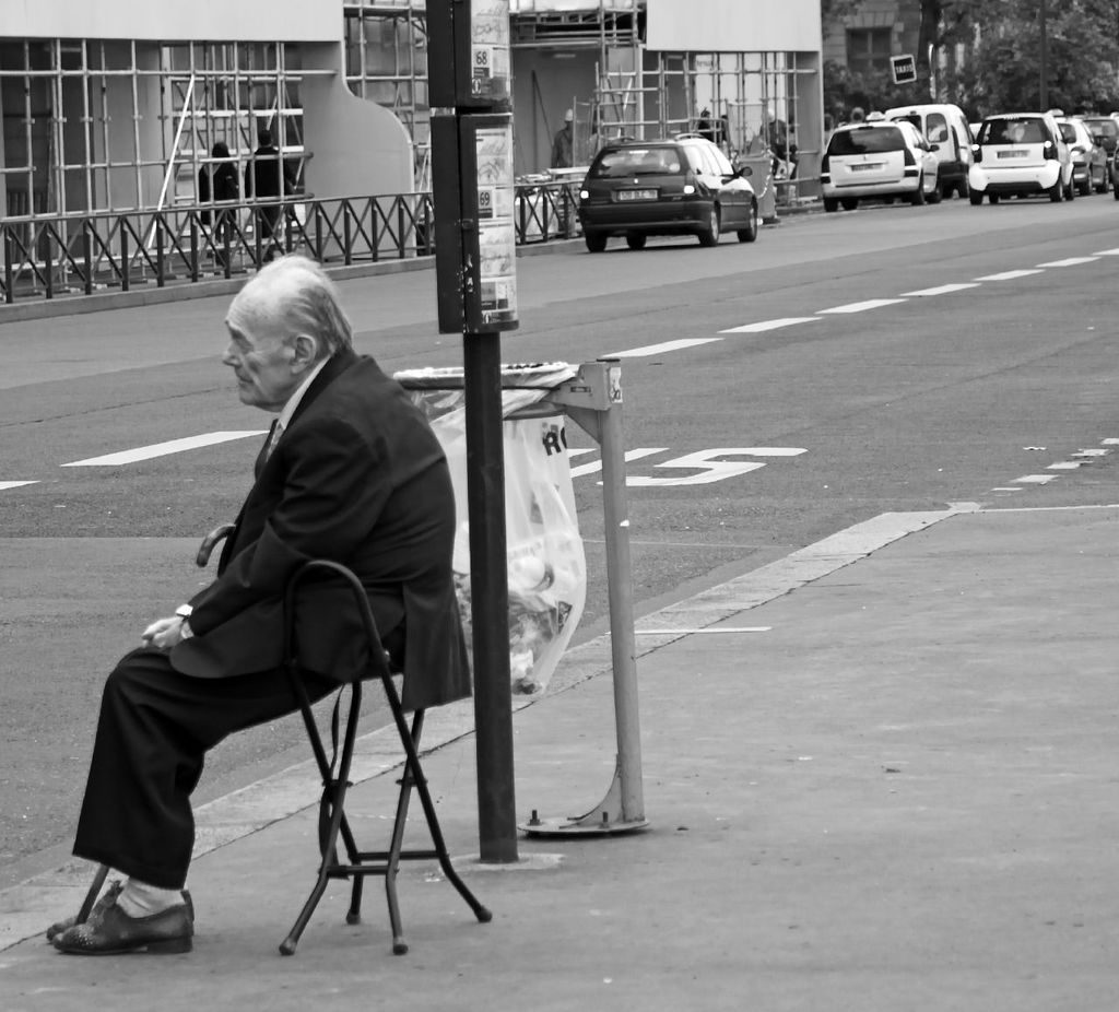 The old man wearing suit is sitting in a chair and there are cars on the road behind him.