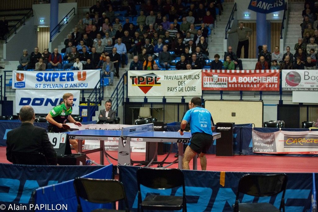 This picture shows a couple of men playing table tennis and we see couple of them watching by...