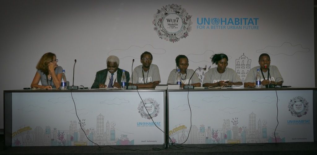 This image is clicked in a conference hall. There are six persons sitting in this image. To the...