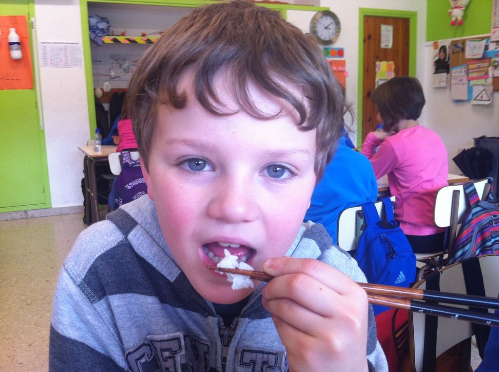 The boy in front of the picture wearing grey and black jacket is eating food with chopsticks....