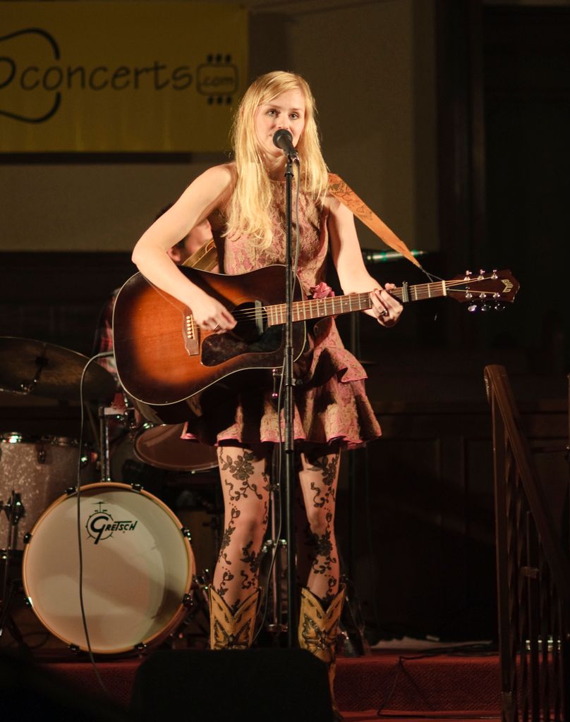This image is clicked in a concert. In the front, there is a woman playing guitar and singing. In...