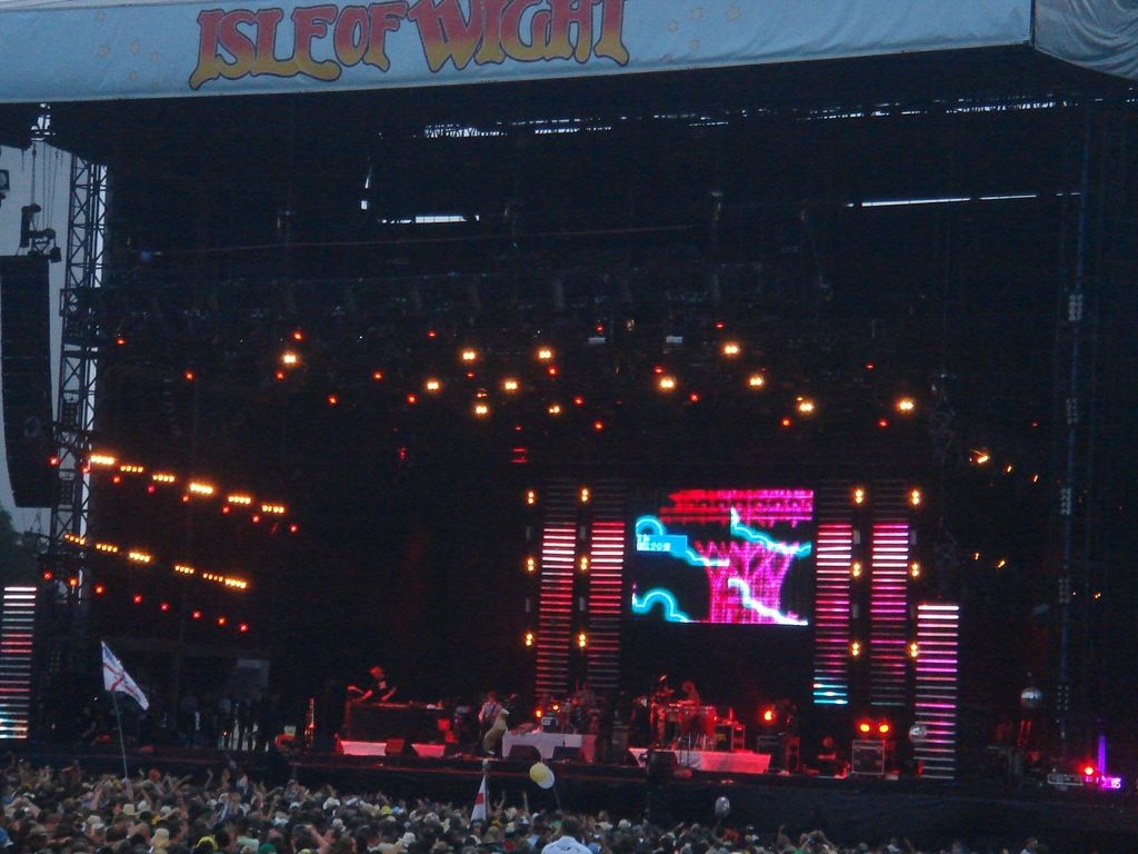 In this image we can see stage with lights. Also there is a screen. And there are people playing...