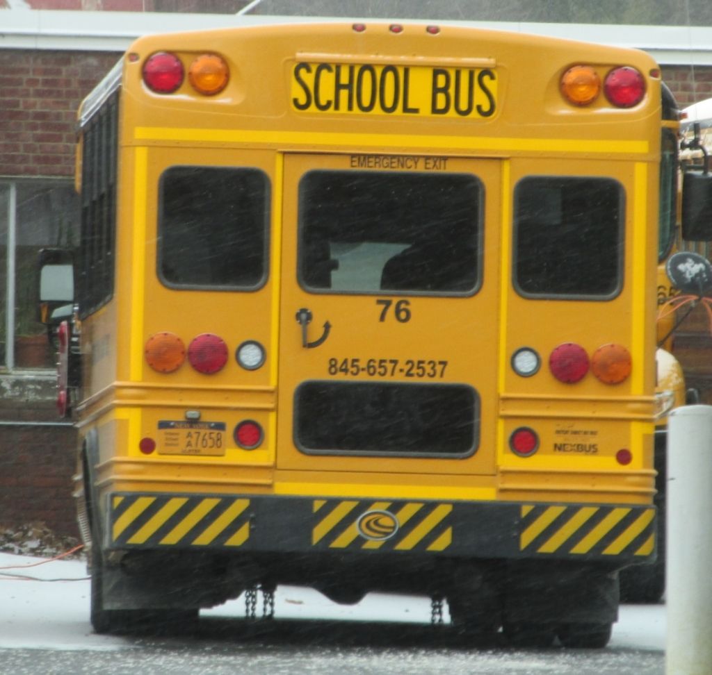 There is a yellow bus on which '' school bus'' is written. Behind that there is a building which...