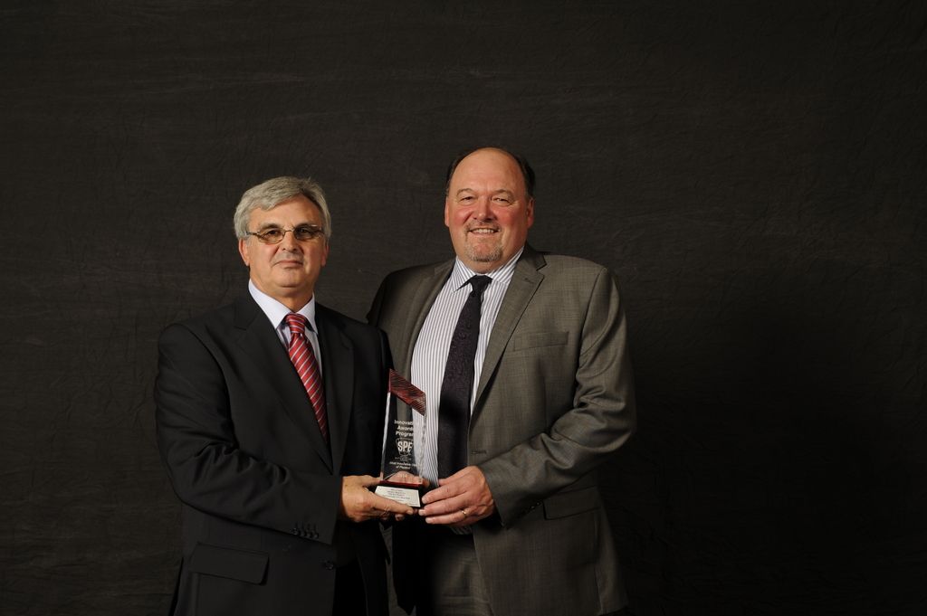 In this image there are two persons wearing suit and tie standing and holding a award in there...