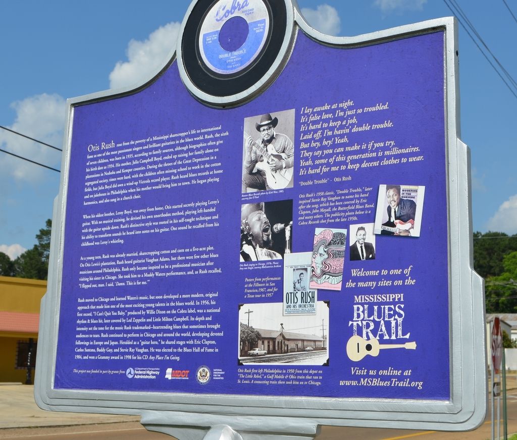 In this image we can see the information board, buildings, shed, trees, electric cables and sky...