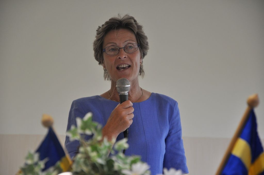 This picture shows a woman standing and speaking with the help of a microphone and we see two Flags...