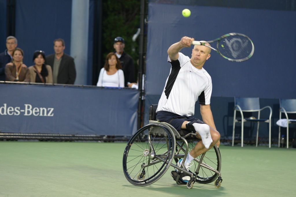 In this image there is a man sitting and running in a wheel chair playing a tennis racket game , at...