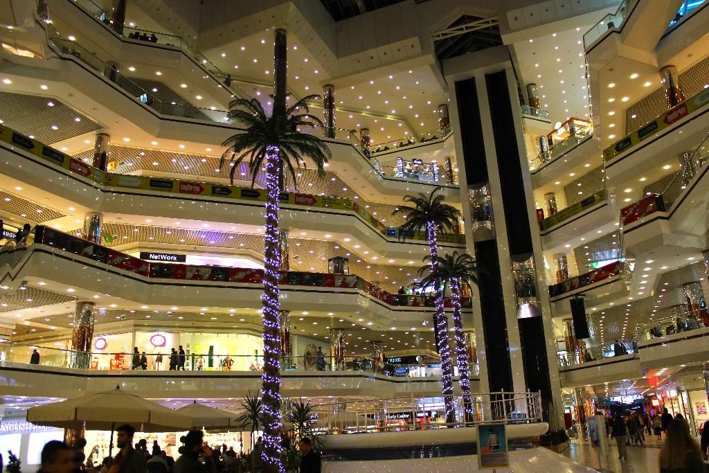 This is an inside view of an shopping complex, where there are trees, umbrellas with the poles,...