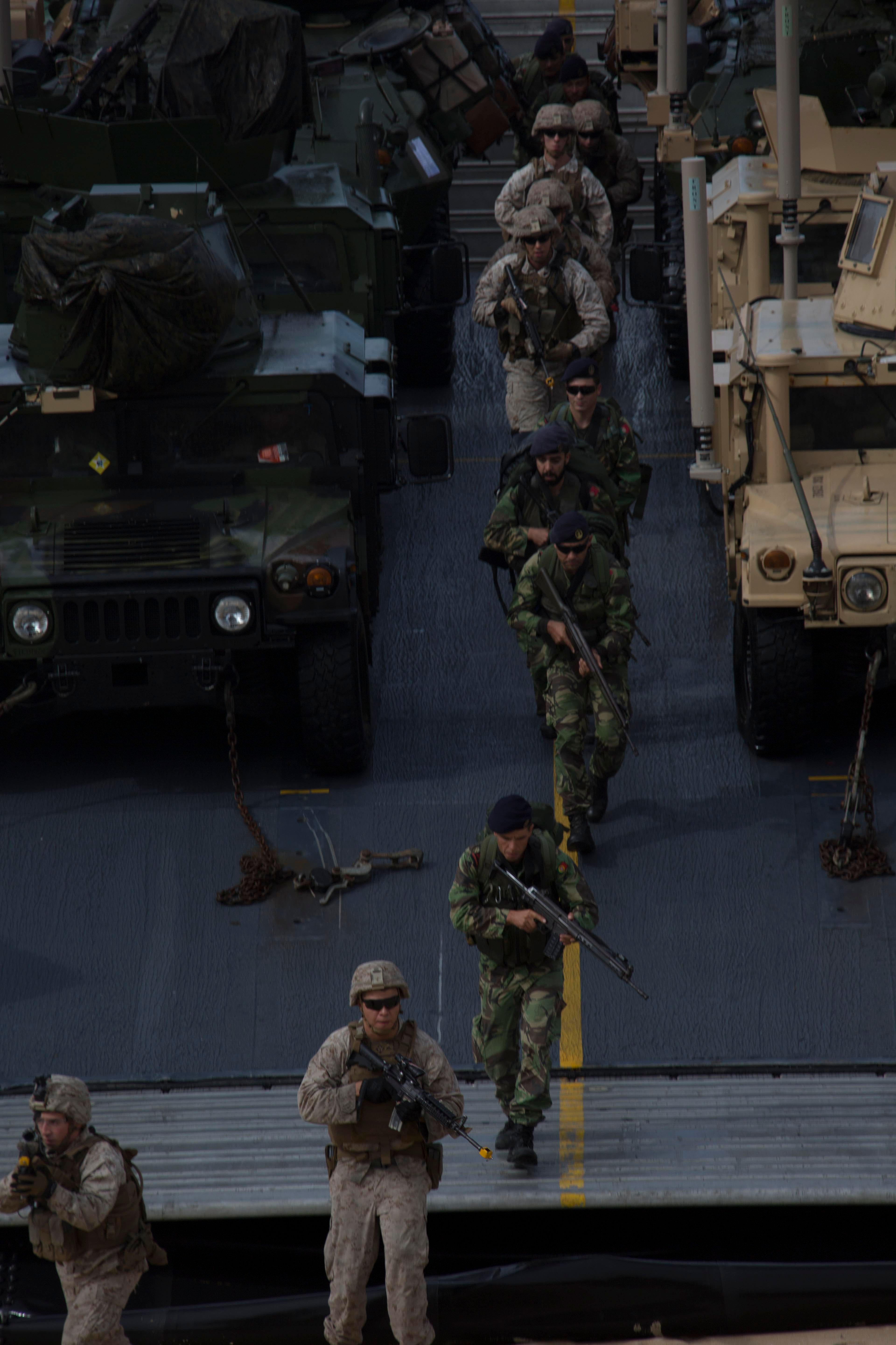The image shows a group of soldiers wearing helmets and holding guns, walking across a bridge with...