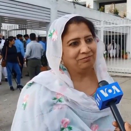 The image shows a woman in a white sari holding a microphone, surrounded by a group of people...