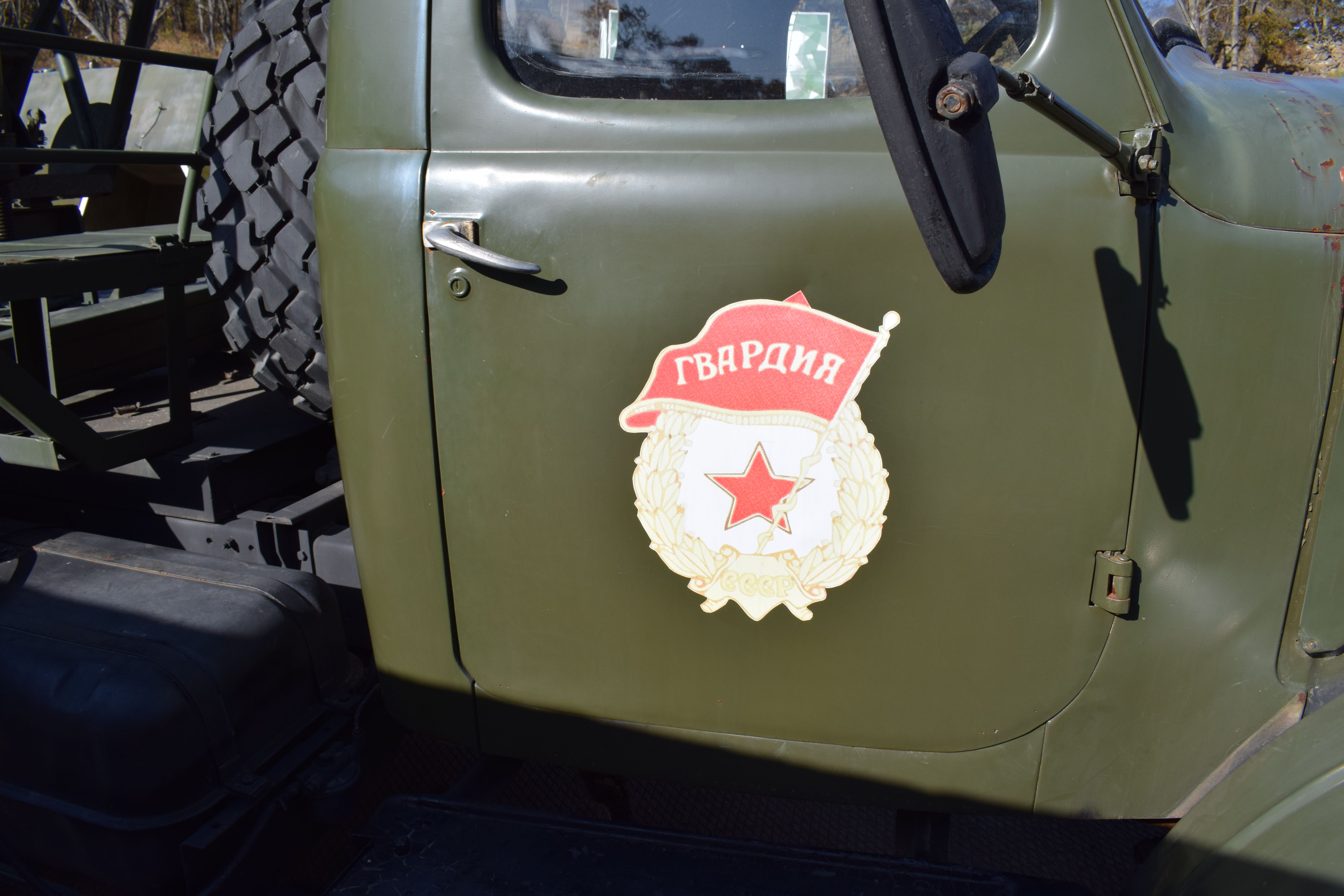 The image shows an old military truck with a badge on the side of it, parked in front of a backdrop...