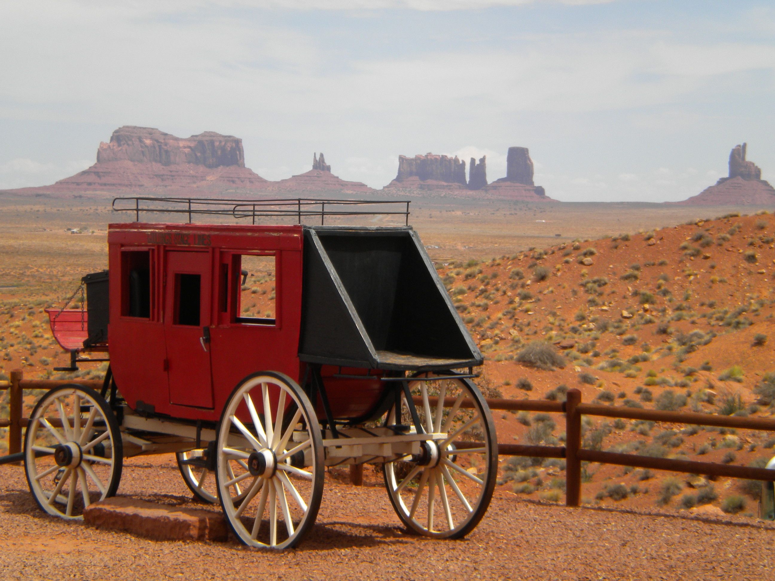 The image shows a stagecoach in Monument Valley, Utah, surrounded by a wooden fence and a few...