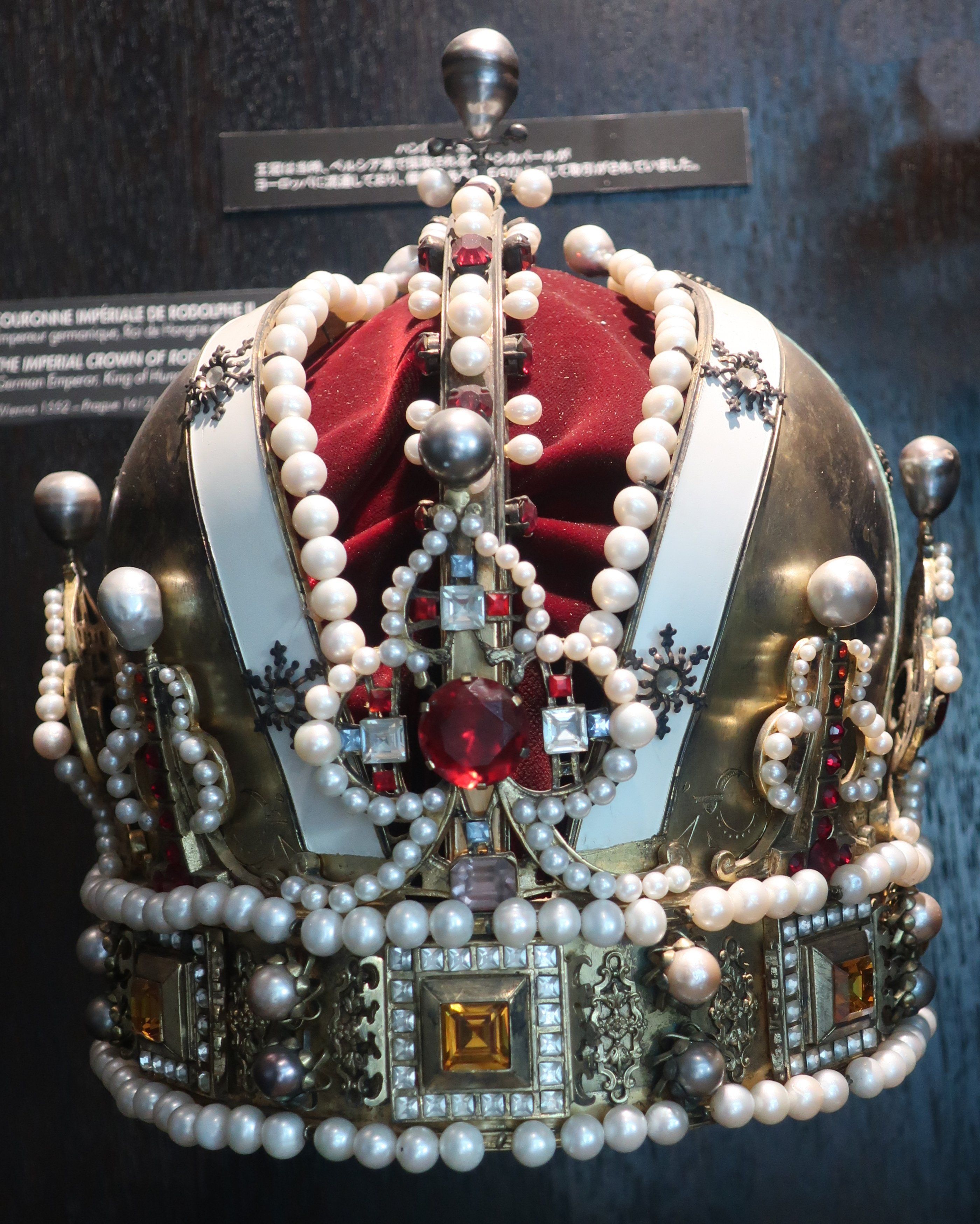 The image shows a gold crown with pearls and jewels on display in a museum, with a board in the...