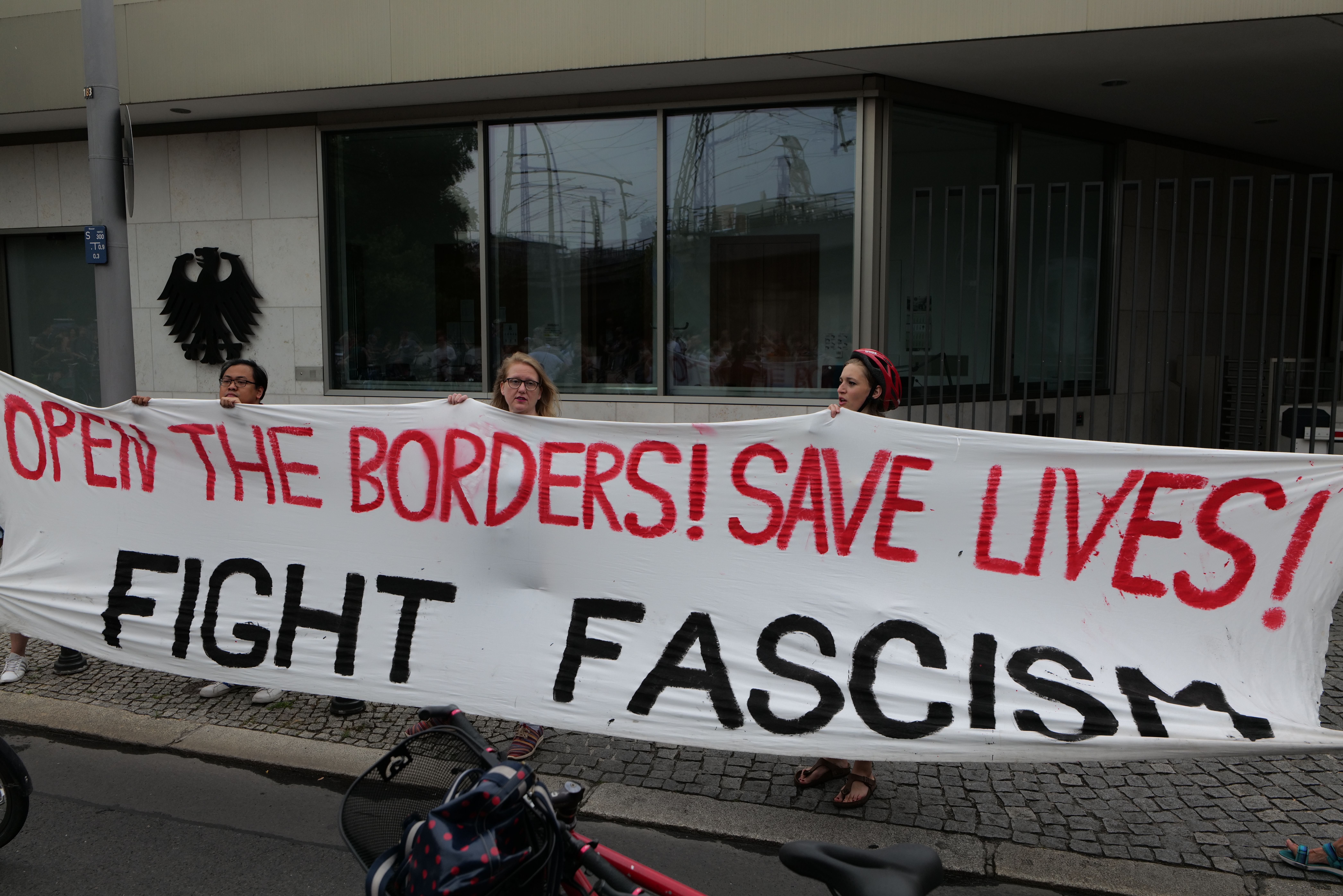 The image shows a group of people holding a banner that reads "Open the Borders, Save Lives, Fight...