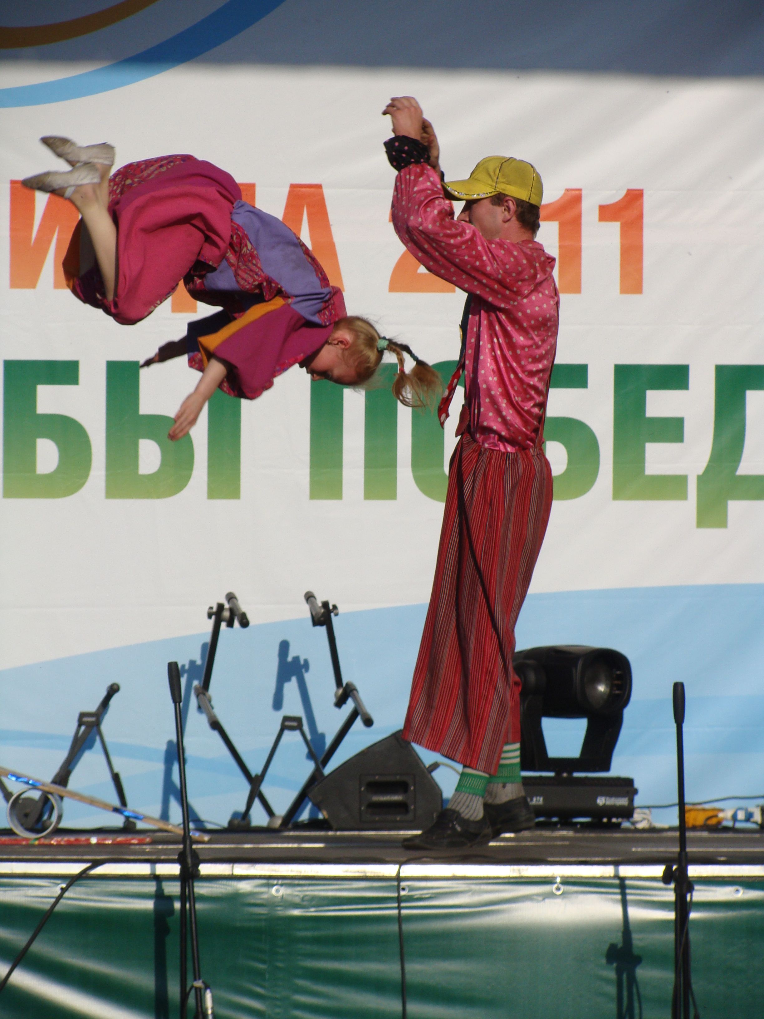 The image shows a man and a woman performing acrobatics on a stage, surrounded by mics with stands,...