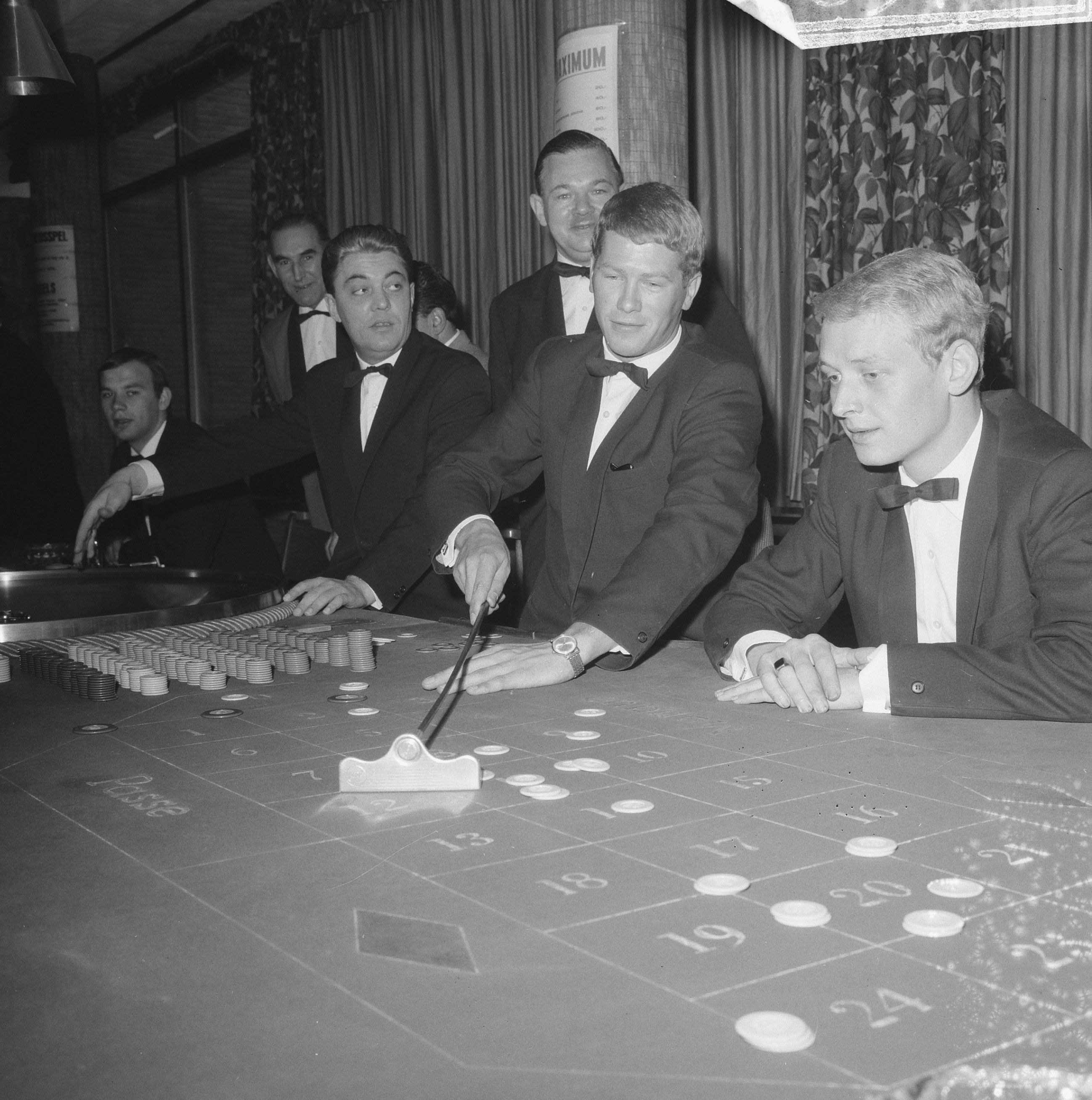 The image shows a group of men in tuxedos playing roulette at a casino table. The table is black...