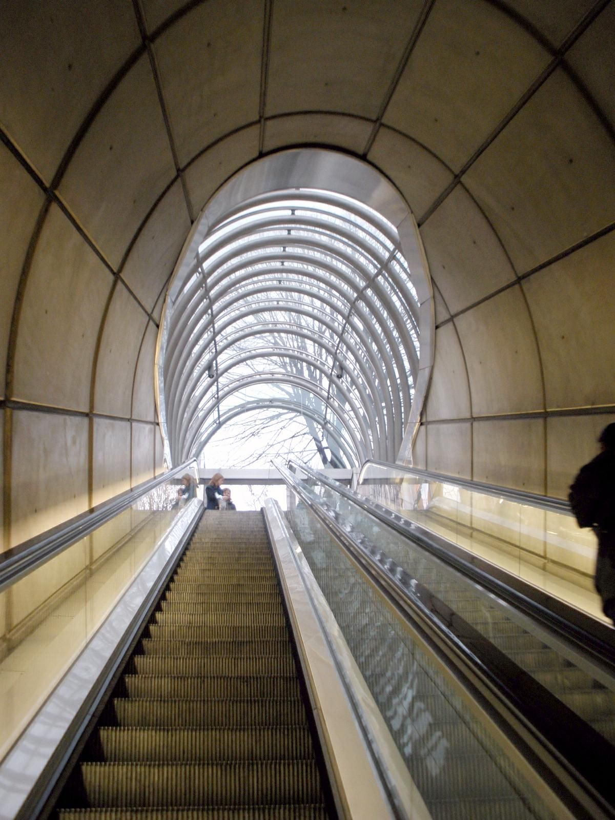 The image shows an escalator in a subway station with people on it, surrounded by walls on either...