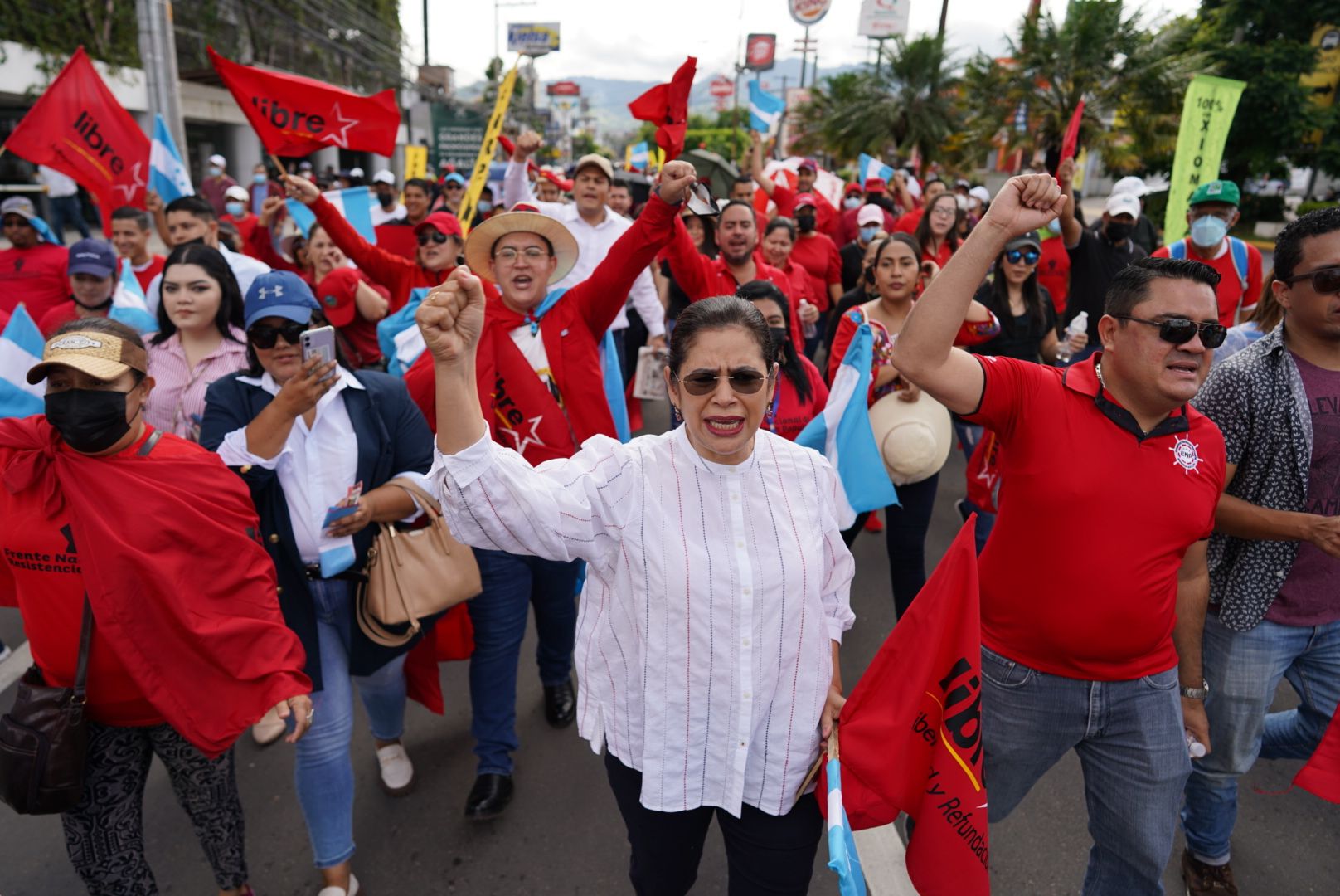 The image shows a large group of people walking down a street, some of them holding flags and...