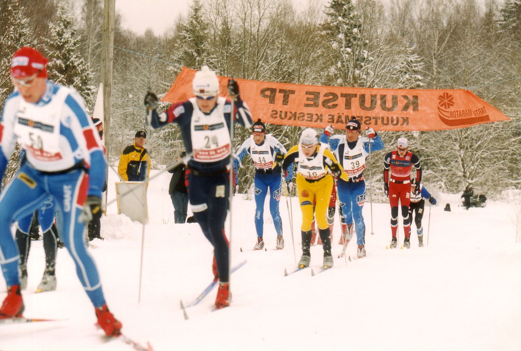 The image shows a group of people skiing down a snow covered slope, wearing caps, goggles, and...