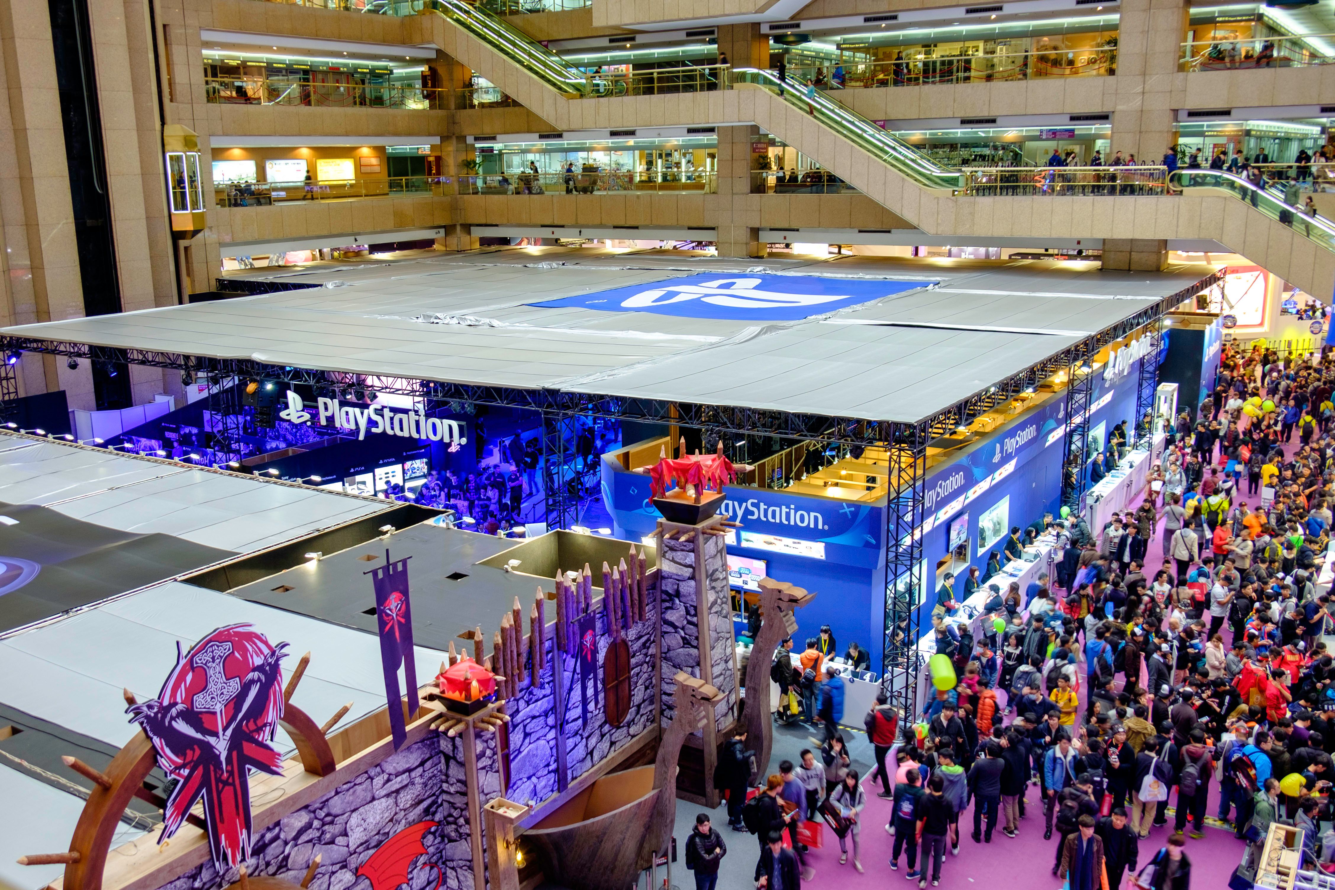 The image shows a large crowd of people standing in a large building with escalators, railings,...