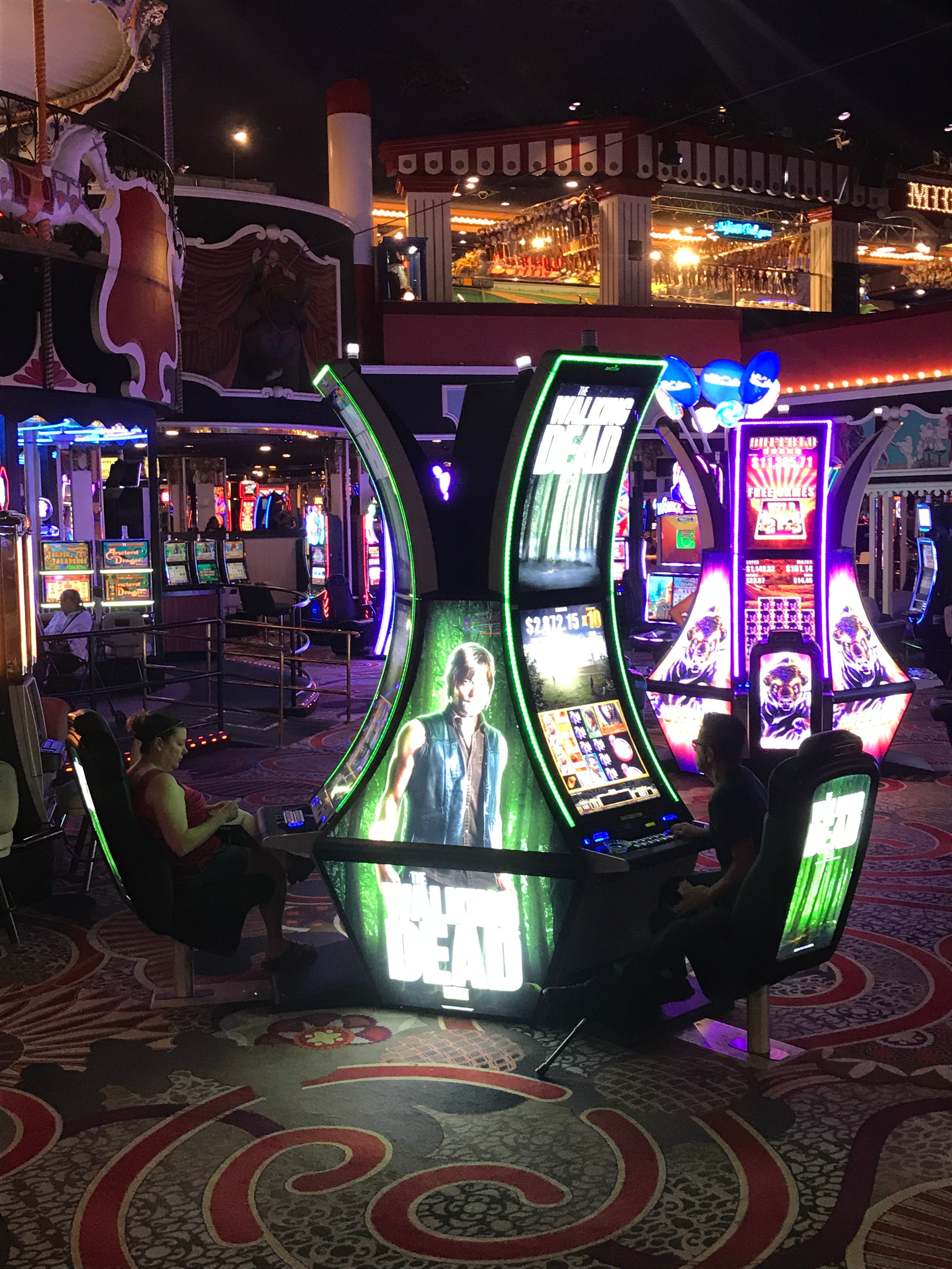 The image shows a group of people playing slot machines in a casino at night, illuminated by the...
