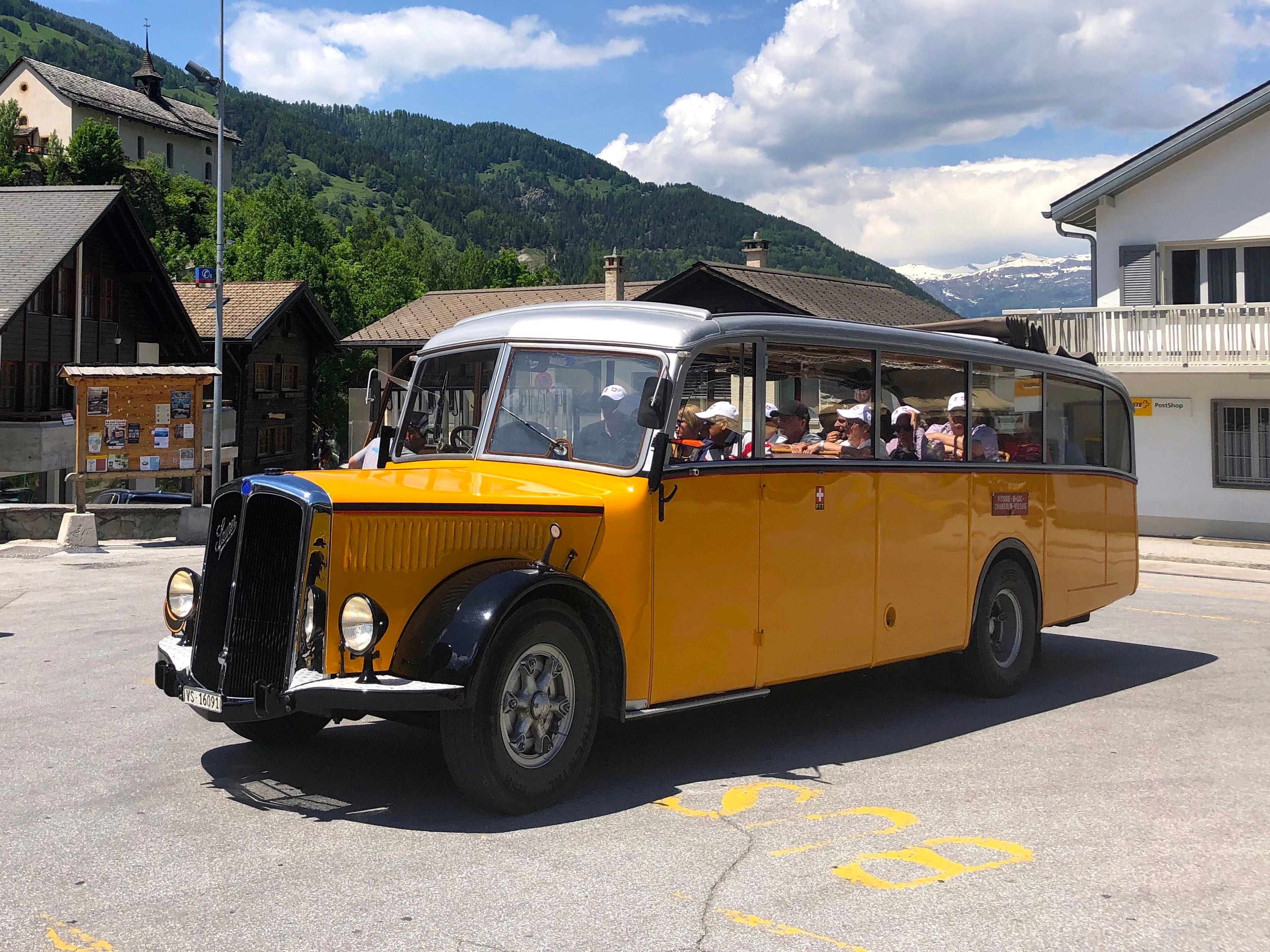The image shows a yellow bus driving down a street lined with houses and trees, with mountains in...