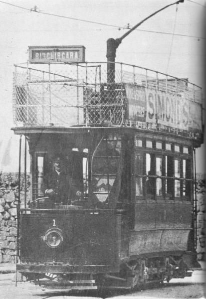 The image shows an old black and white photo of a tram on the tracks, with a few people inside the...