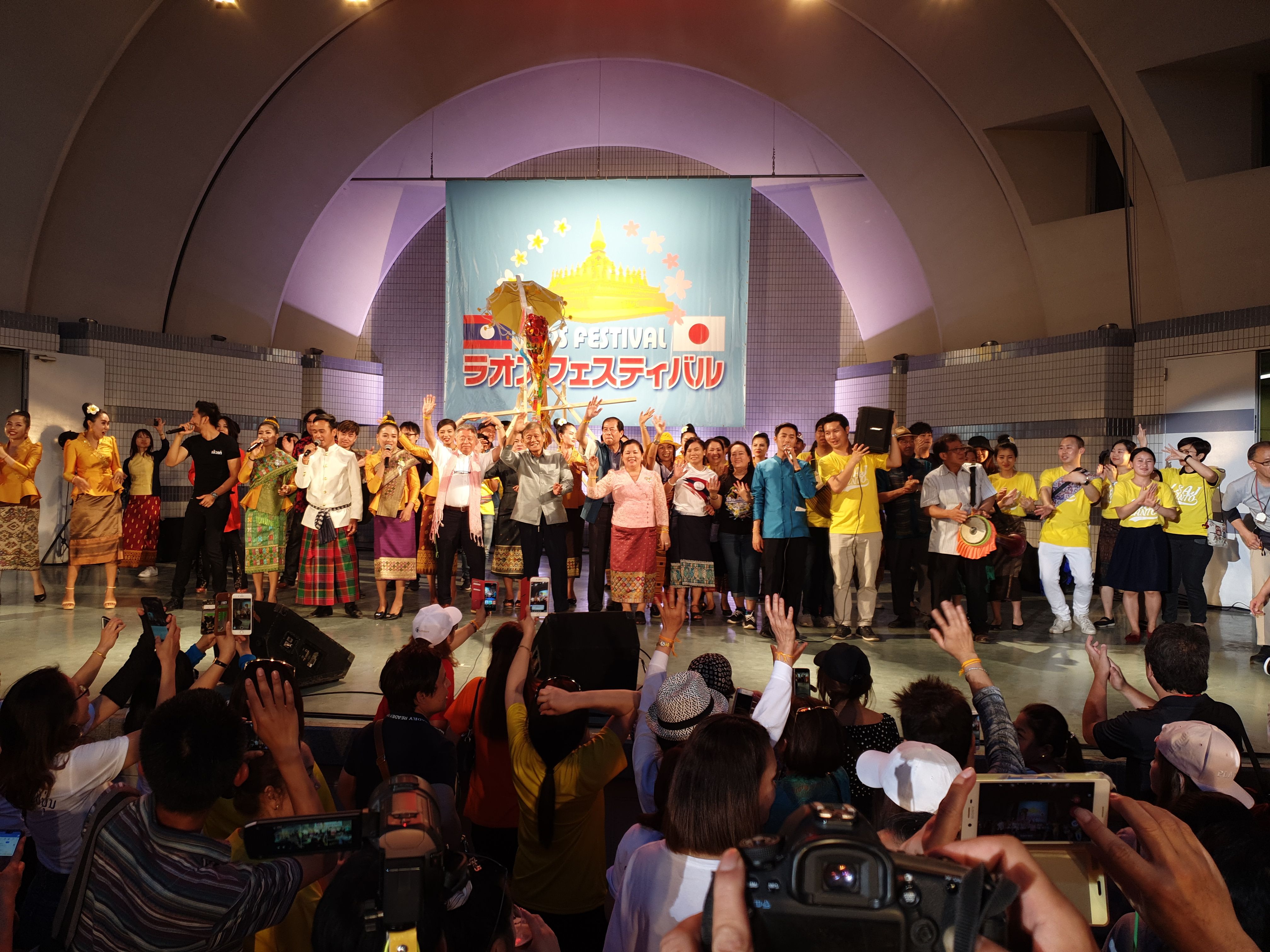 The image shows a group of people standing on top of a stage, some of them holding cameras and...