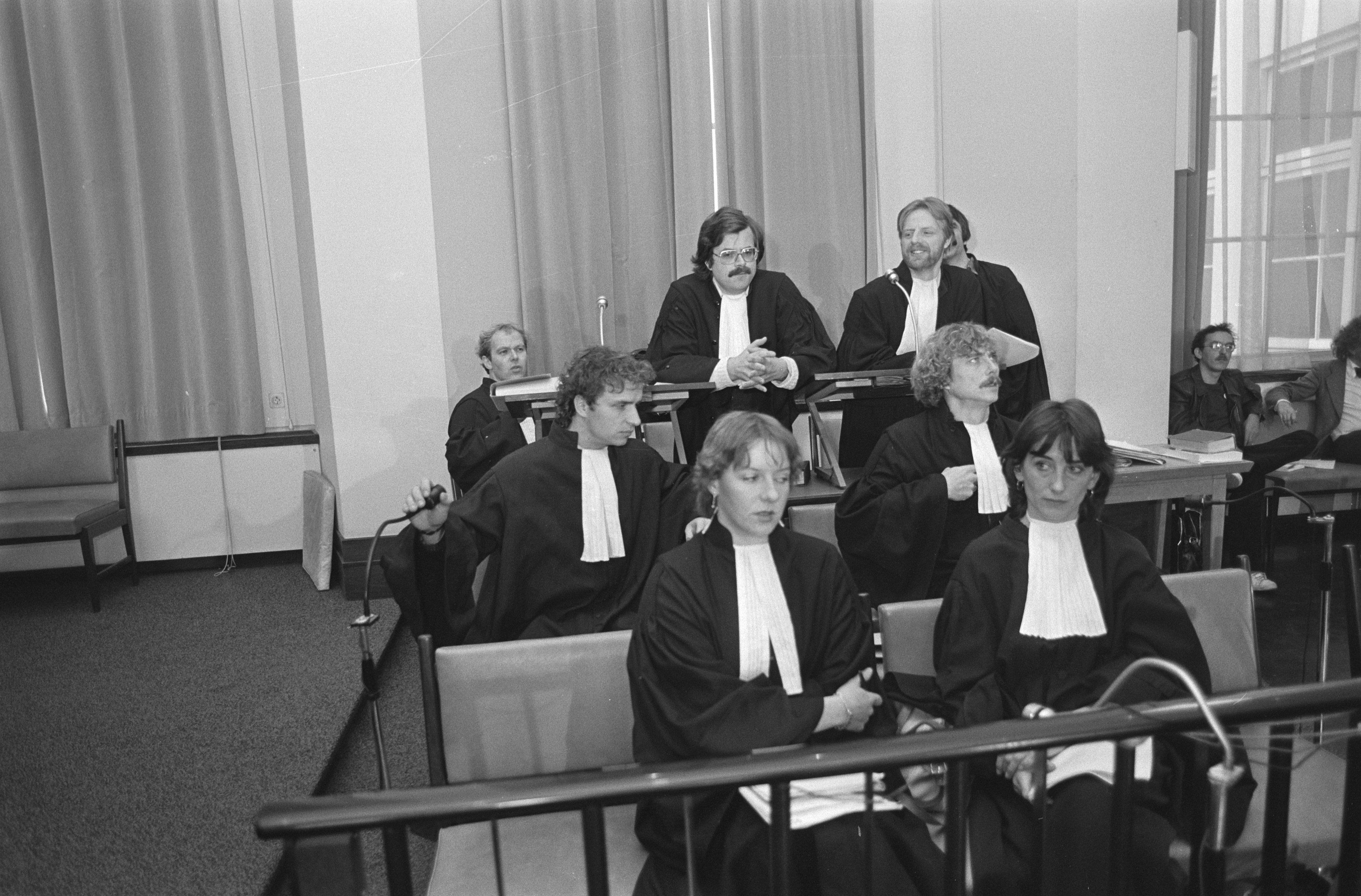 The image shows a group of people sitting in a courtroom, with a railing in the foreground and a...