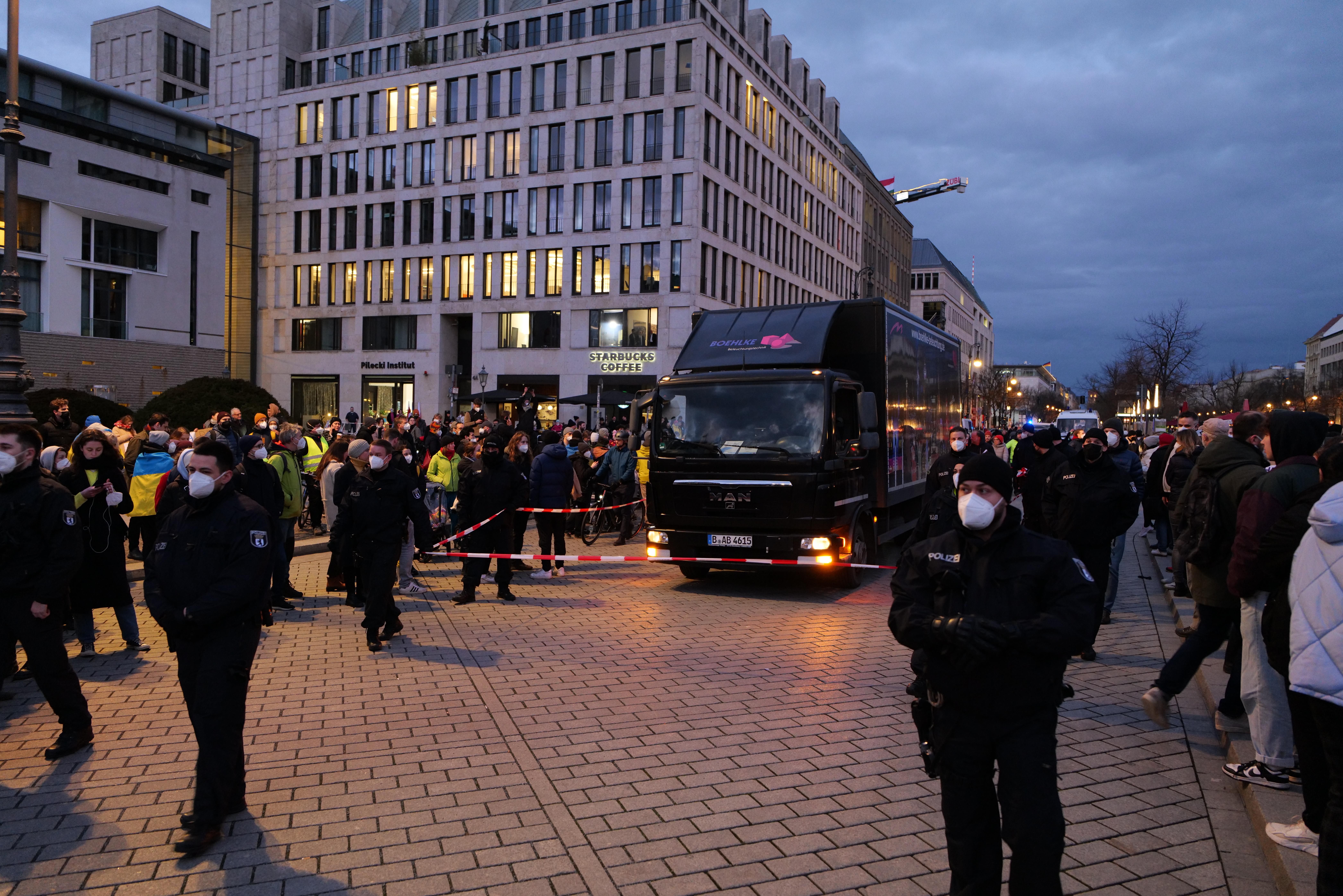 The image shows a group of people standing in front of a truck on a road surrounded by buildings...