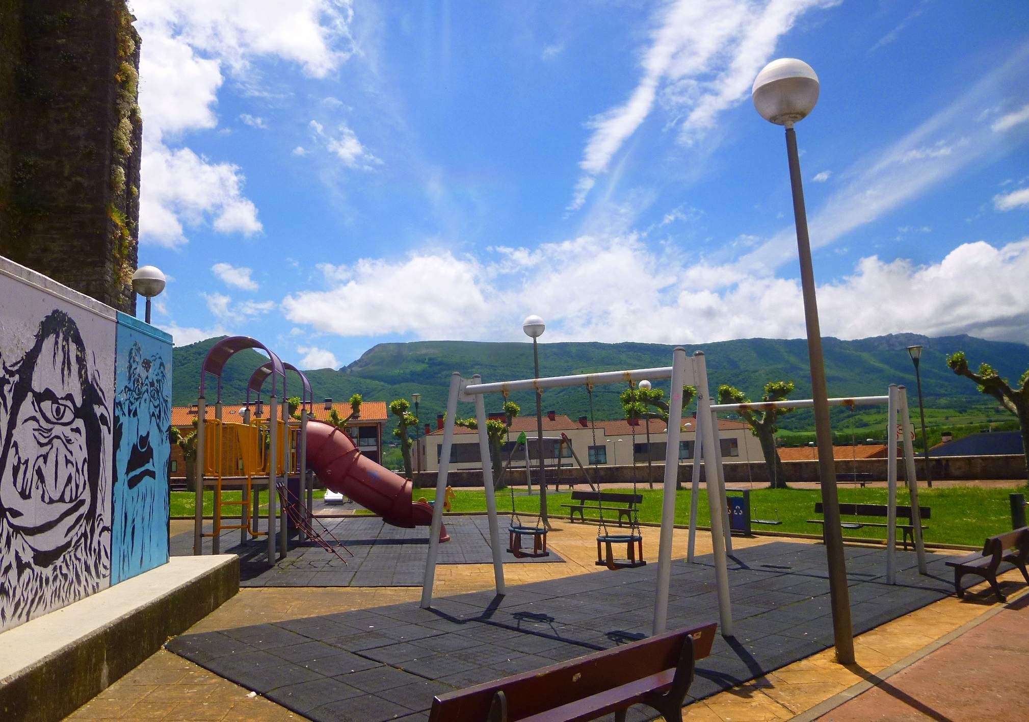 The image shows a playground with swings, a slide, benches, poles, lights, a wall with a painting,...
