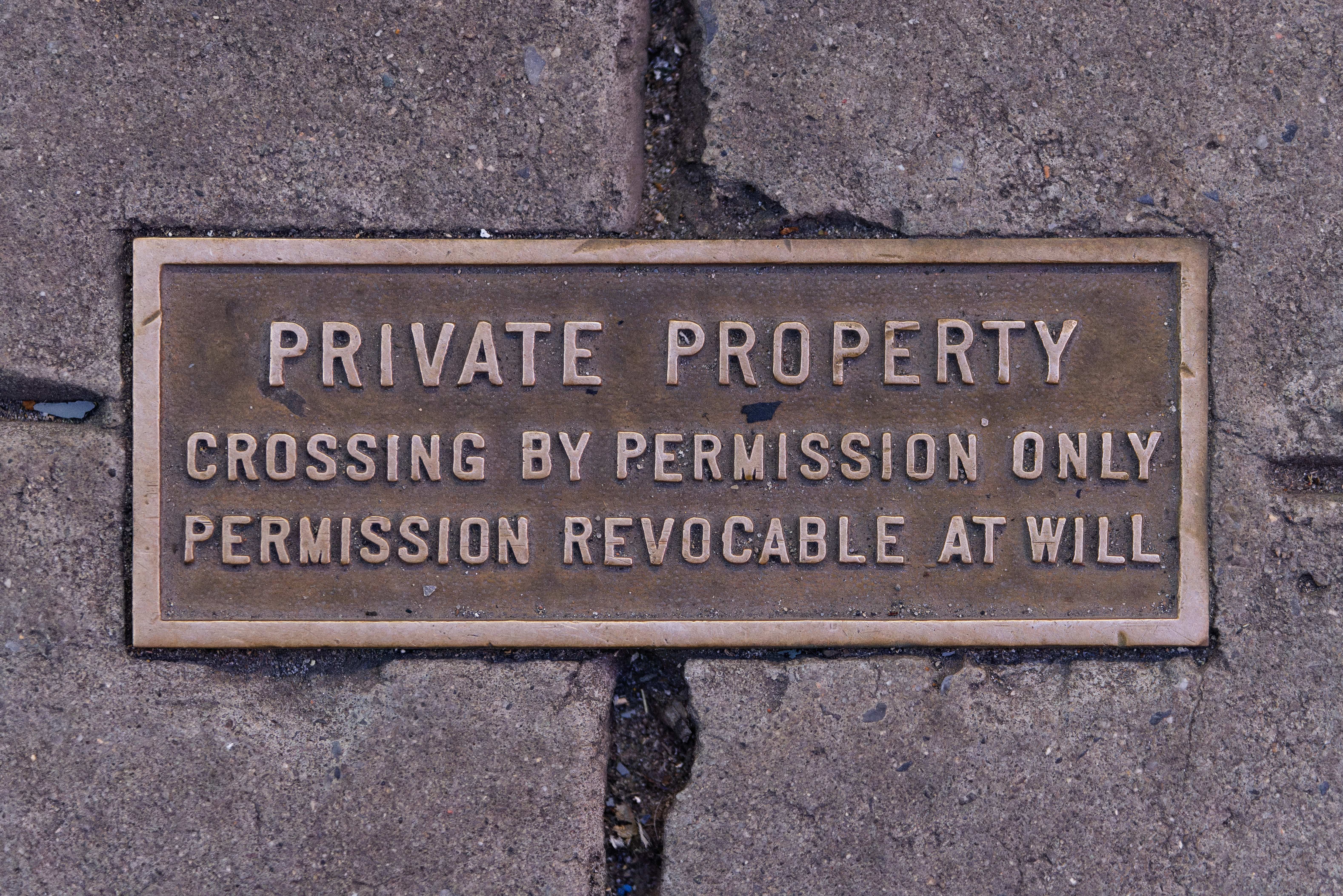 The image shows a plaque on the sidewalk that reads "Private Property Crossing by Permission Only"....