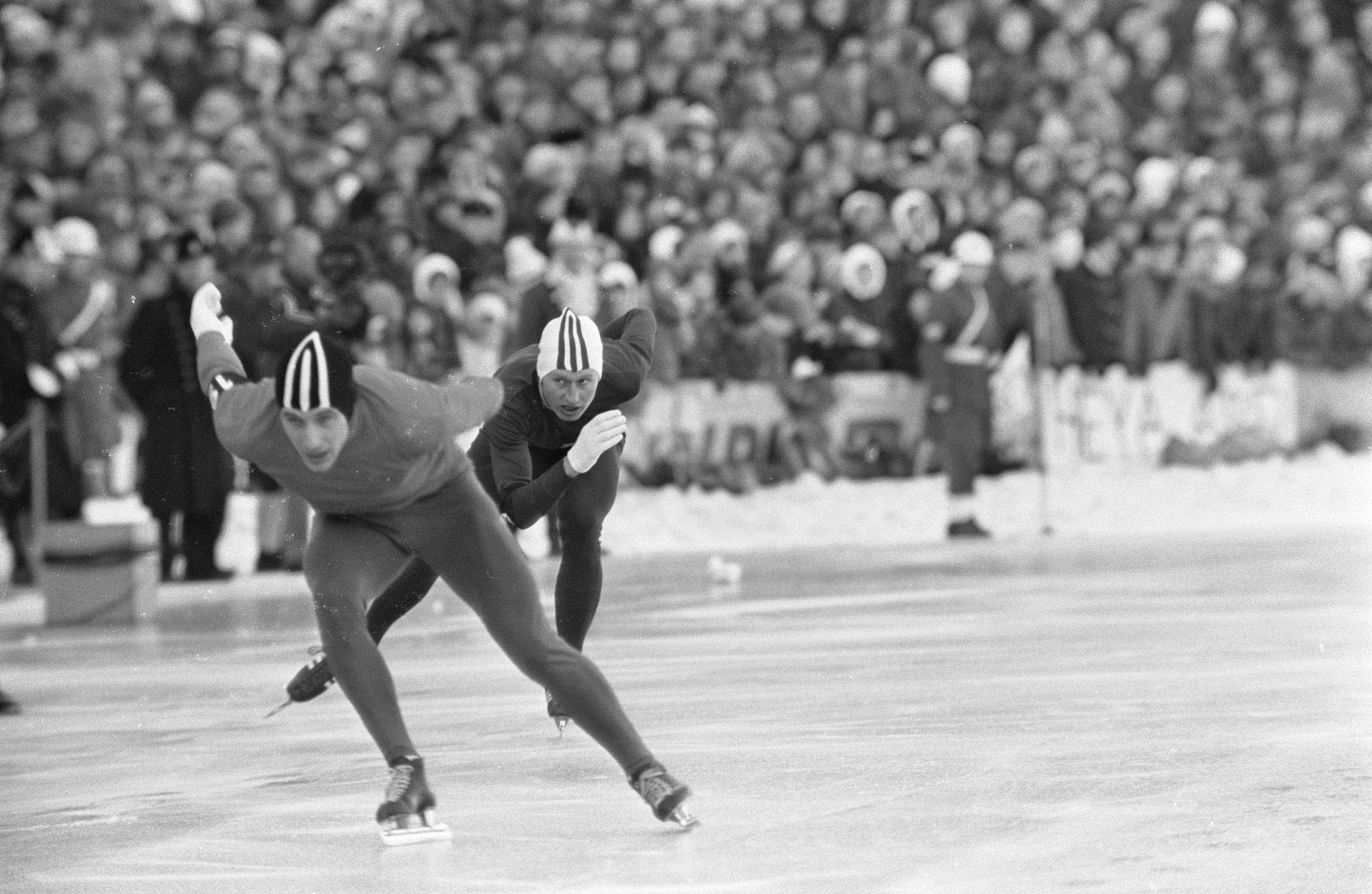 The image shows two men speed skating on an ice rink in front of a large crowd. They are both...
