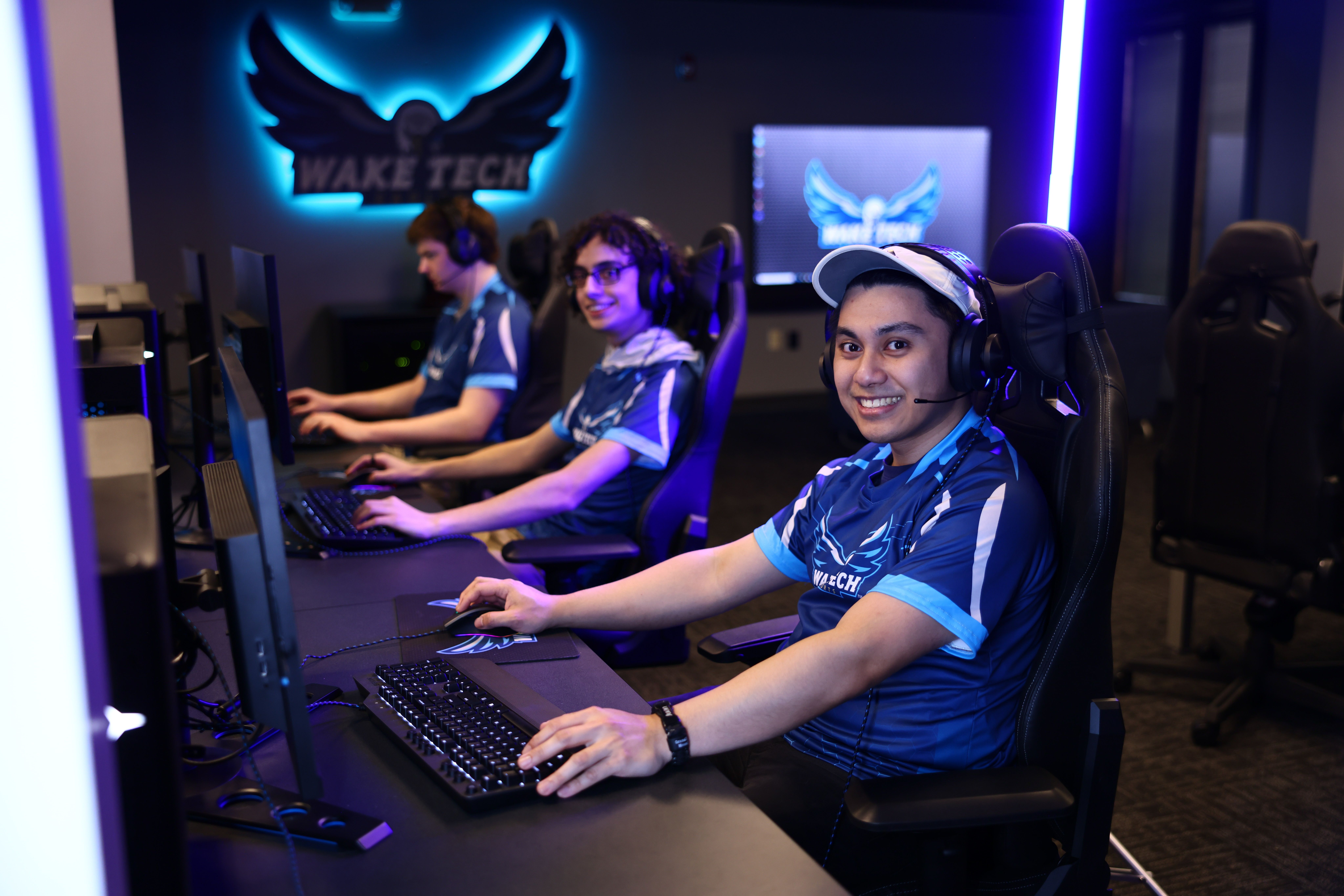 The image shows a group of people sitting at a desk in front of computers, wearing headsets and...