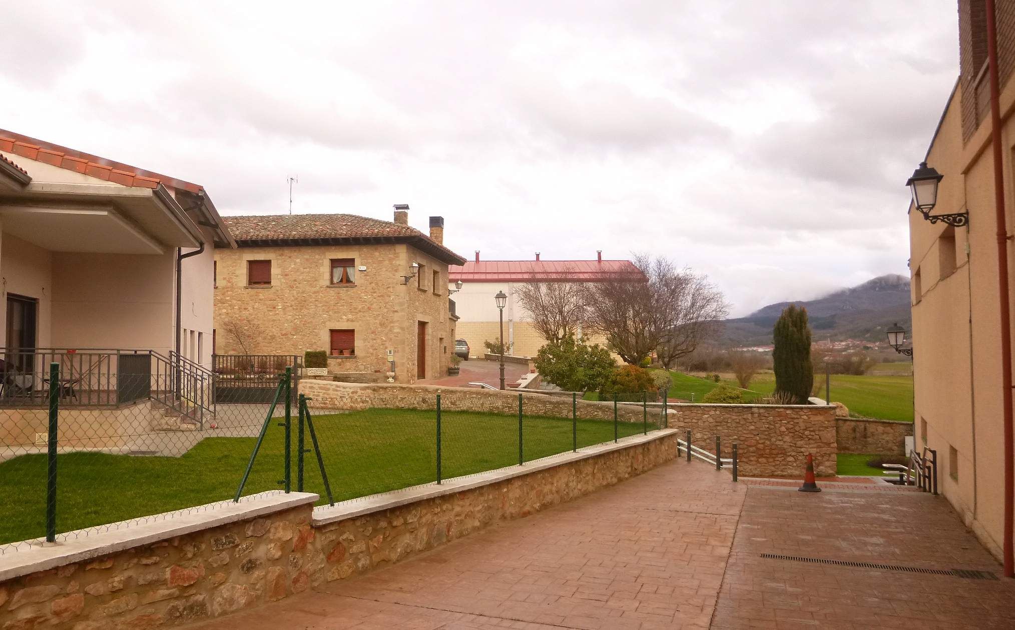 The image shows a building with windows, railings, steps and a roof, surrounded by a metal fence,...