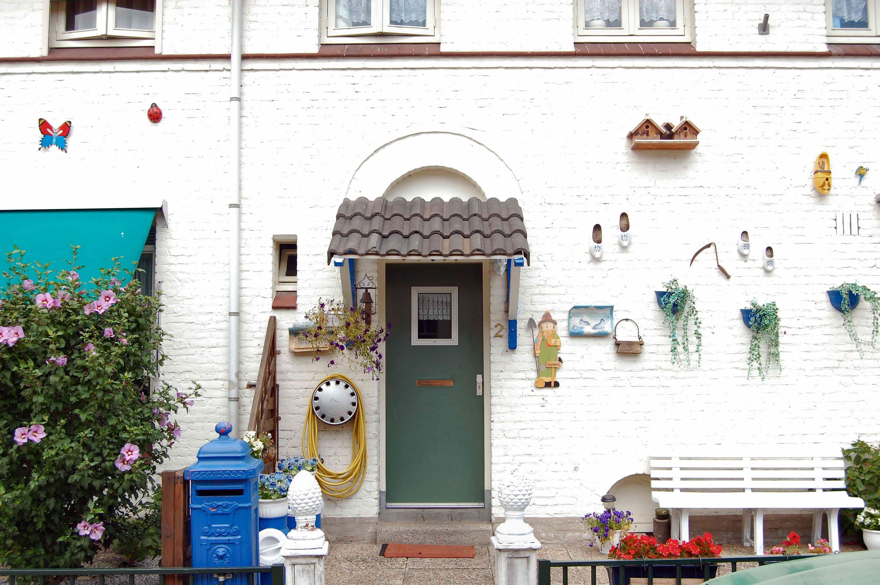 The image shows a white house with a green door and windows, a bench in front of it, plants with...