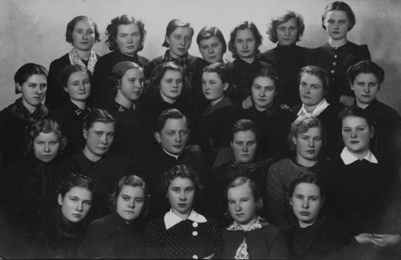 The image shows a black and white photo of a group of women in school uniforms, with a wall in the...