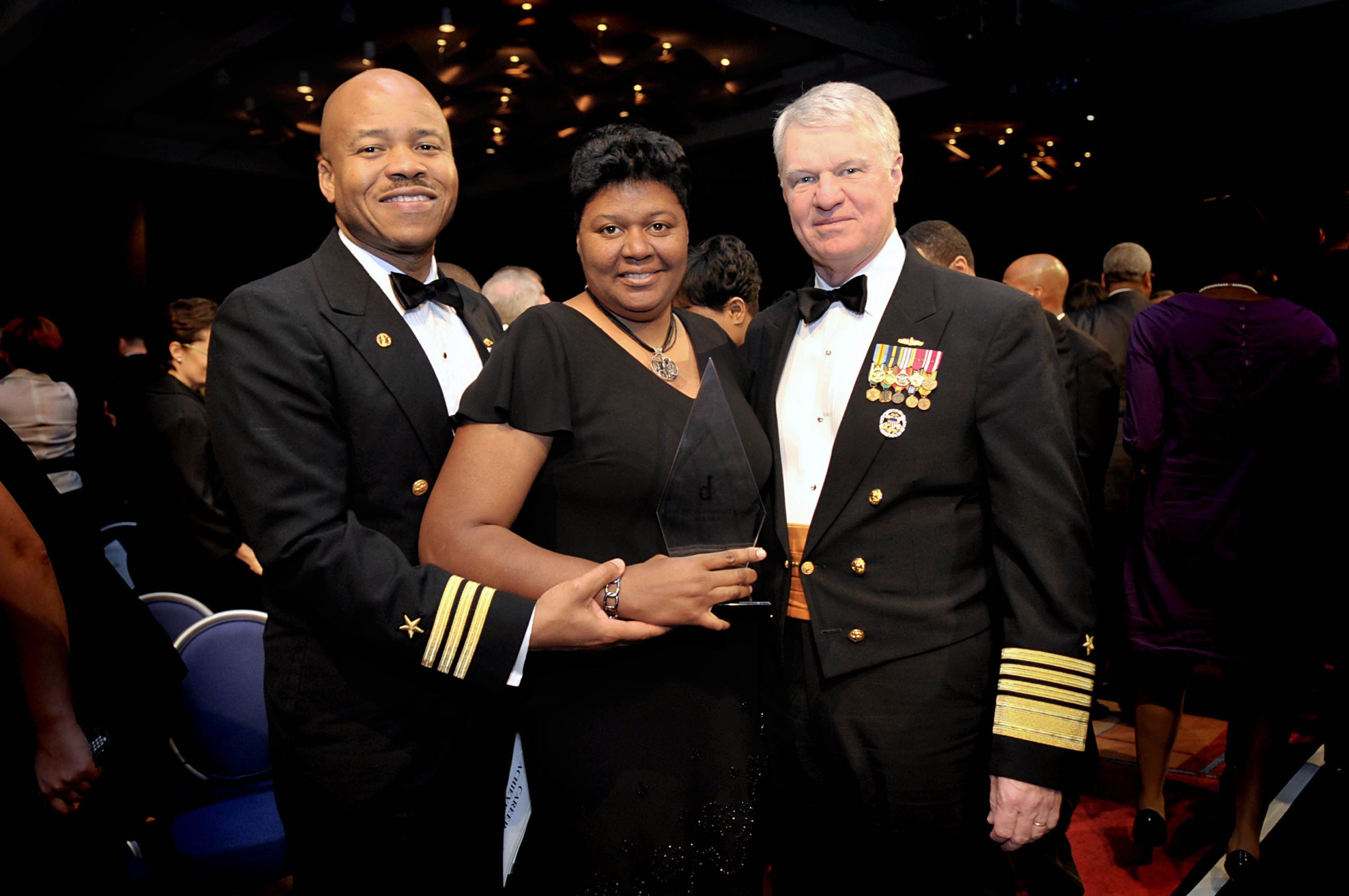 The image shows two men and a woman standing next to each other on a red carpet. The woman is...