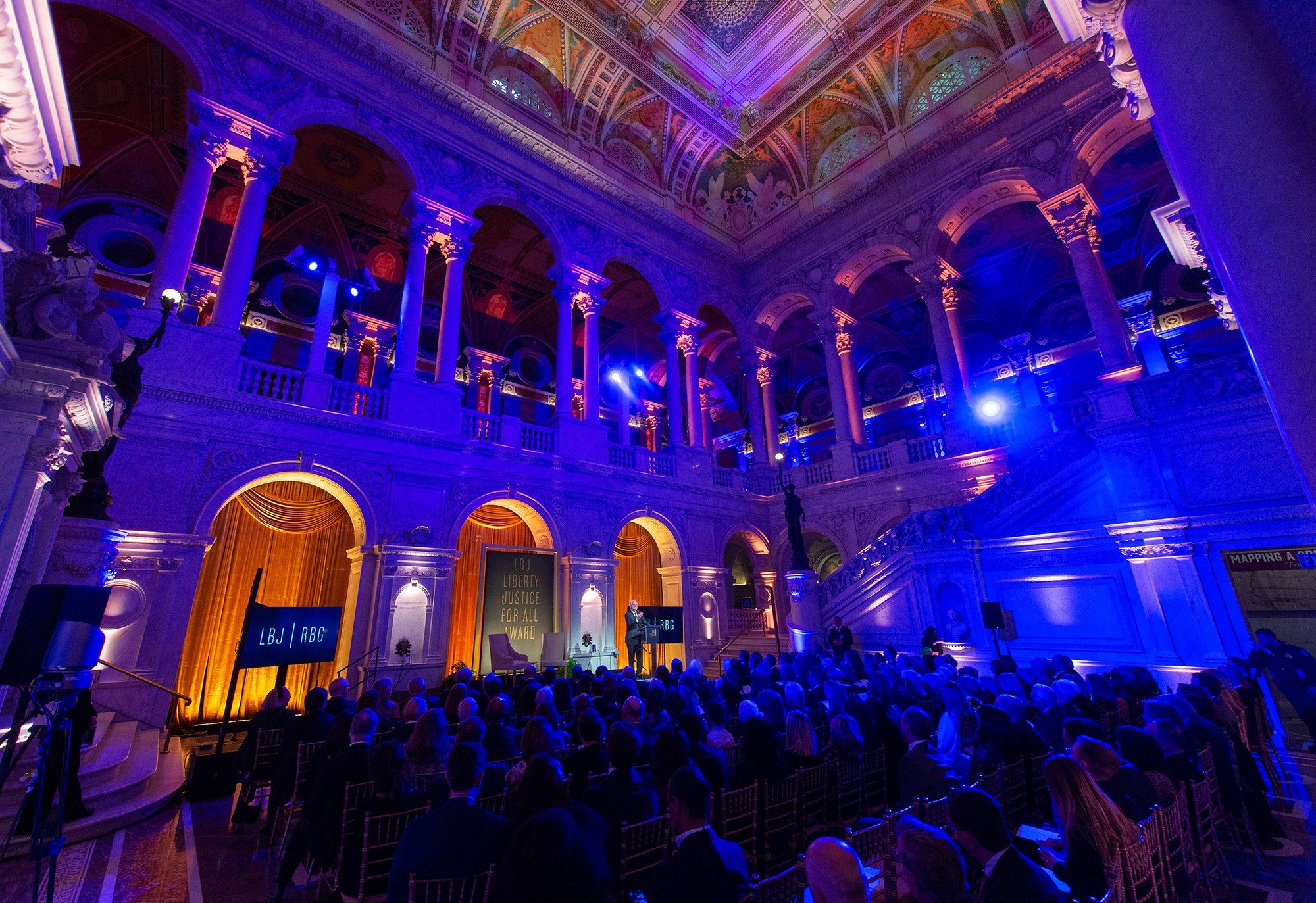 The image shows a large group of people sitting in chairs in a large room with a stage in the...