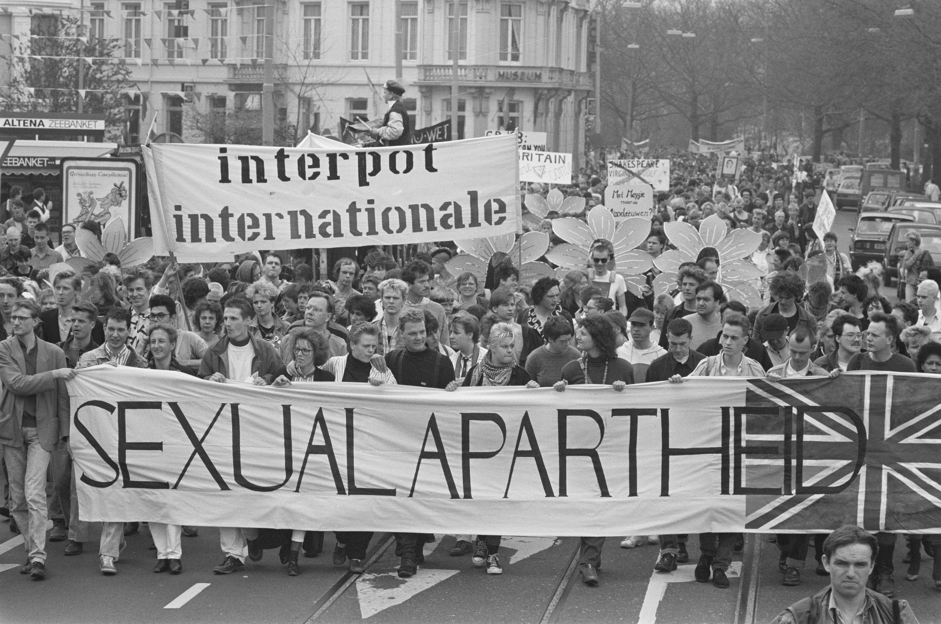 The image shows a group of people walking down a street, holding a banner that reads "Sexual...