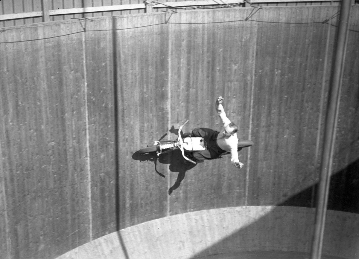The image shows a man on a motorcycle doing a trick in the air, with a wall in the background and a...