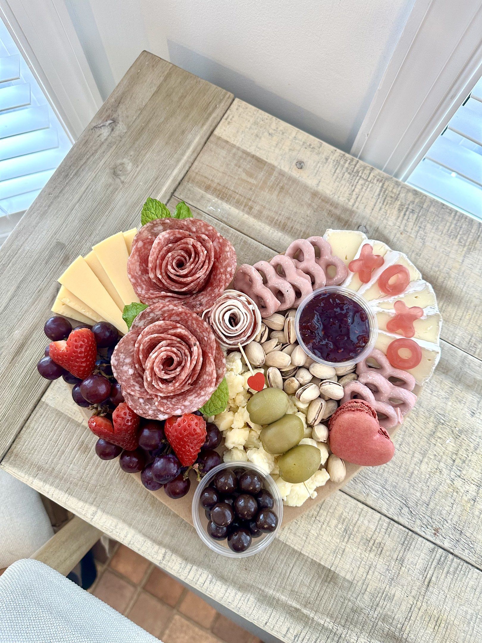 The image shows a wooden table with a heart-shaped platter filled with a variety of cheeses,...