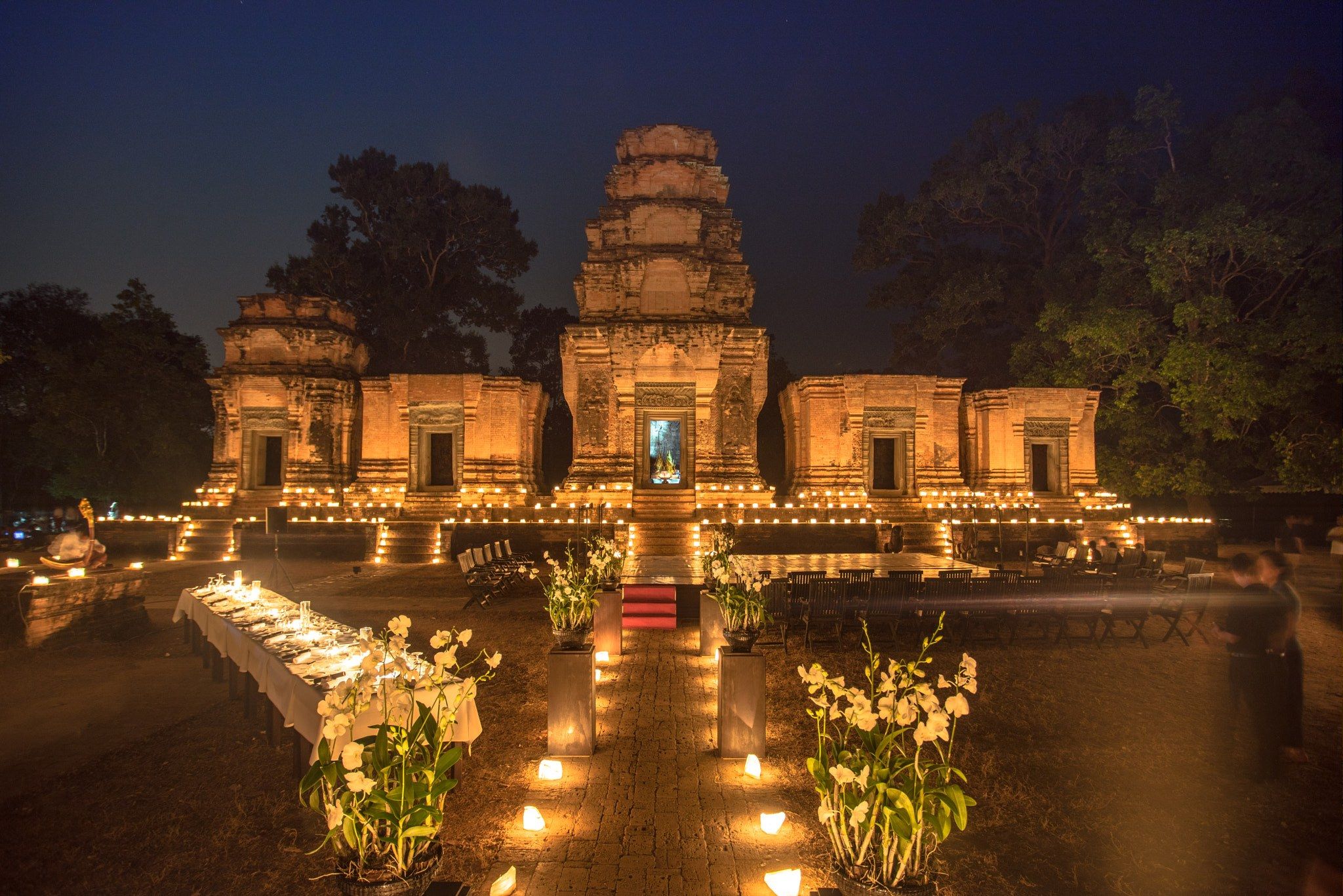 The image shows a group of people standing in front of a temple at night, illuminated by lights and...