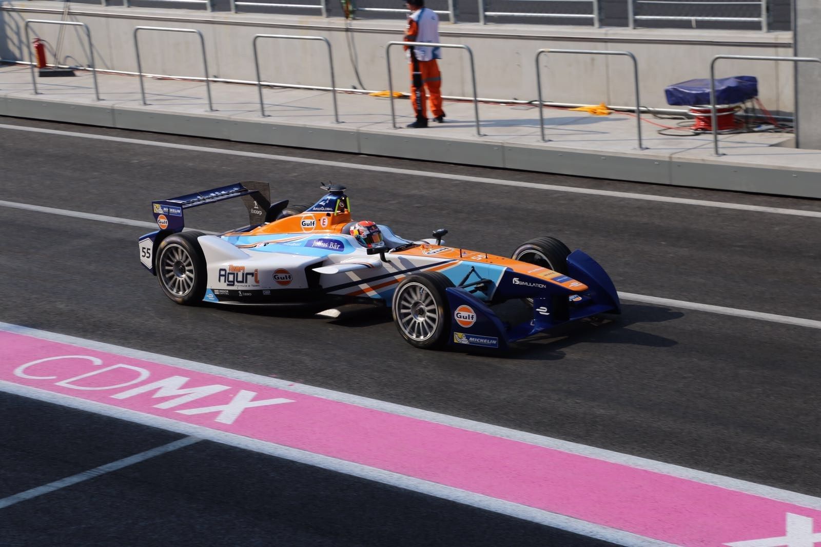 The image shows a man driving a Formula car on a race track, with a person wearing a helmet in the...