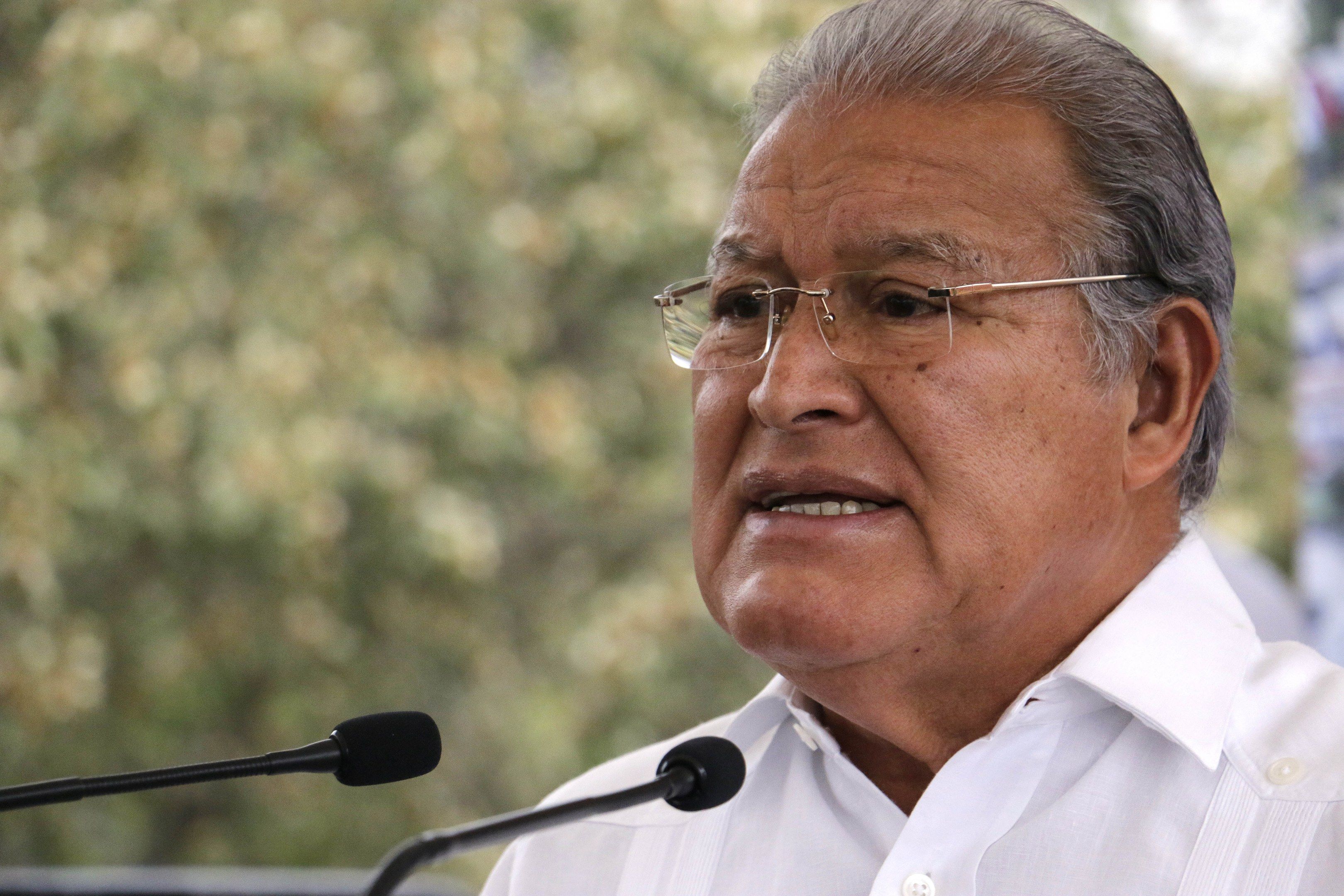 The image shows a man in a white shirt and glasses speaking into a microphone in front of a blurred...