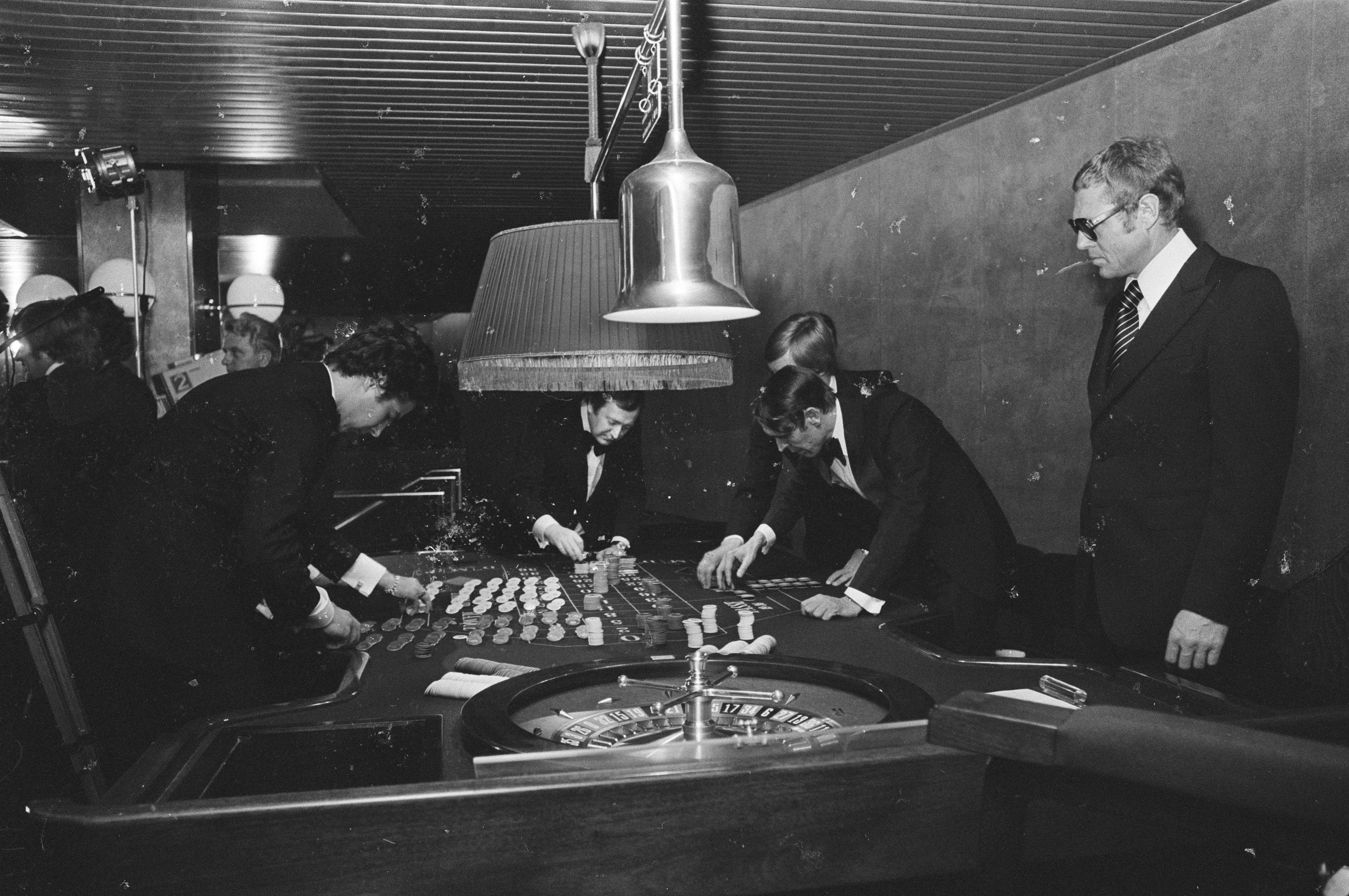 The image shows a group of men playing roulette in a casino. They are wearing blazers, ties, and...